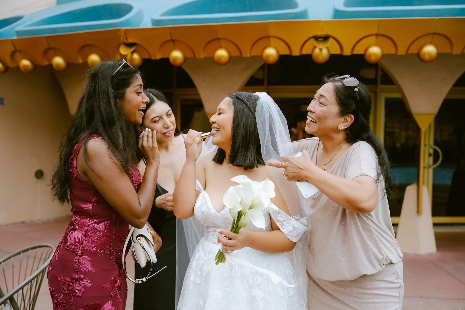 Bride in white gown holds bouquet while bridesmaids apply makeup outdoors at a wedding venue.