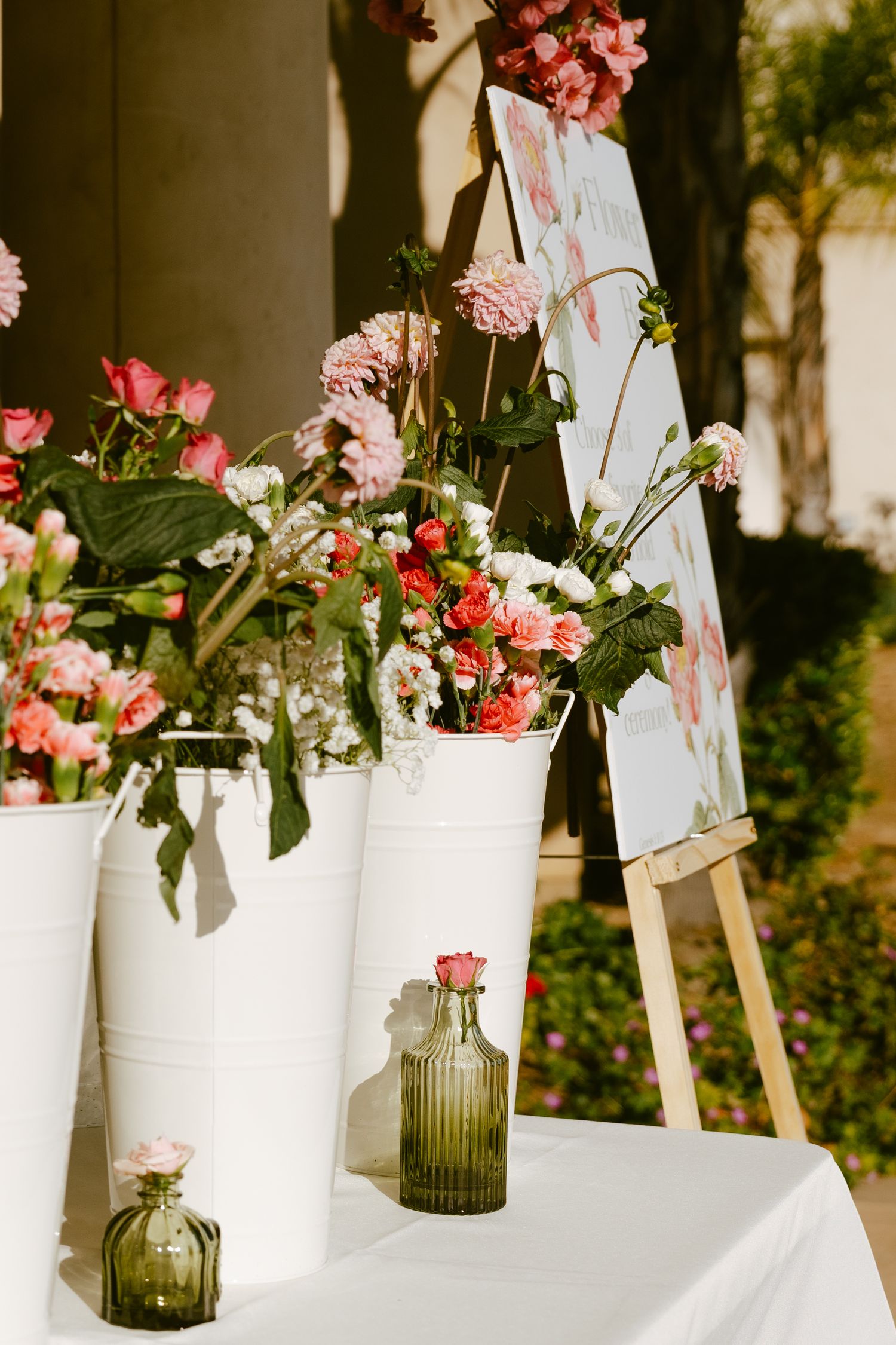 White buckets and green vases filled with pink flowers arranged on a white table next to a wooden easel with signage.