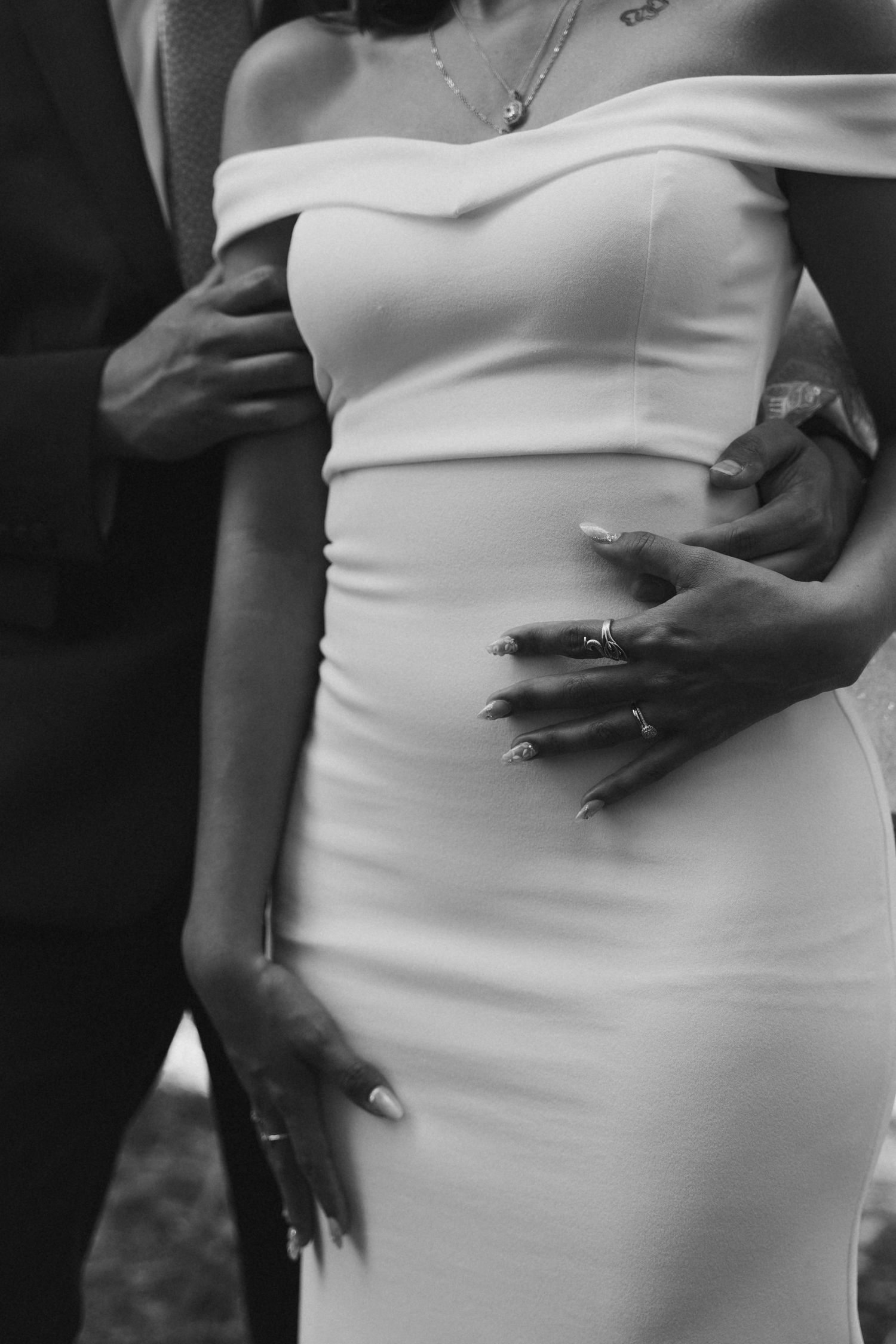Close-up black and white photo of bride in off-shoulder white gown with grooms hands on her waist, showing wedding rings.