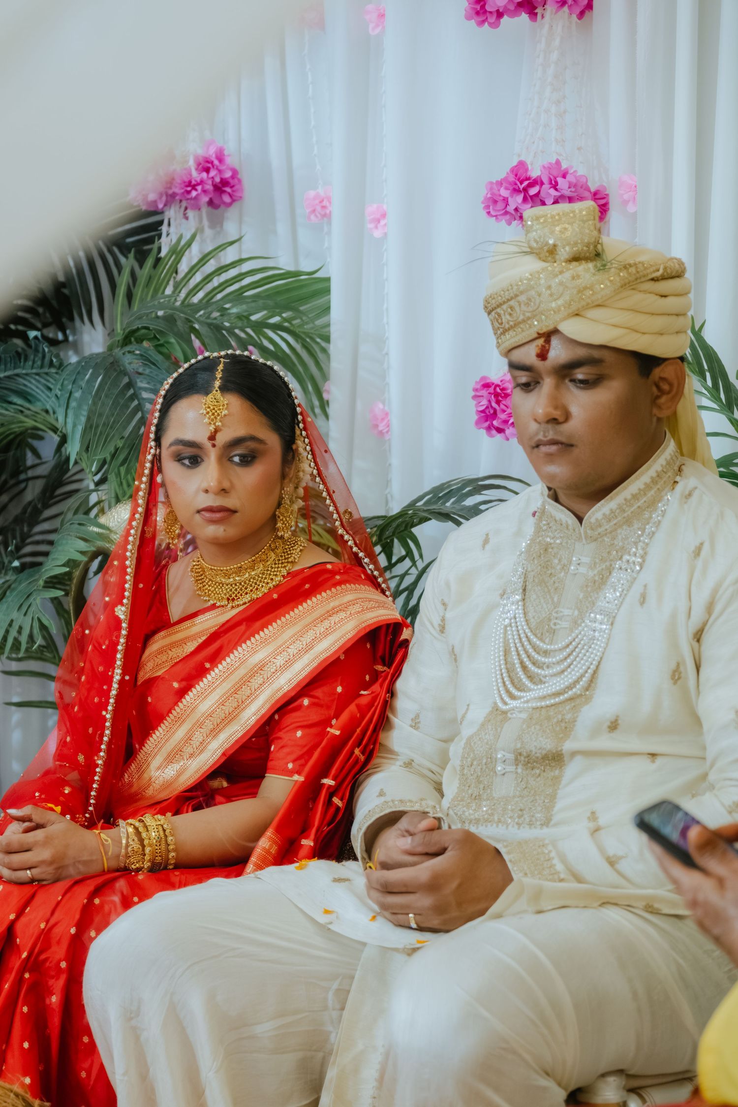 Indian bride in red saree and groom in white sherwani and gold turban seated during traditional Hindu wedding ceremony.