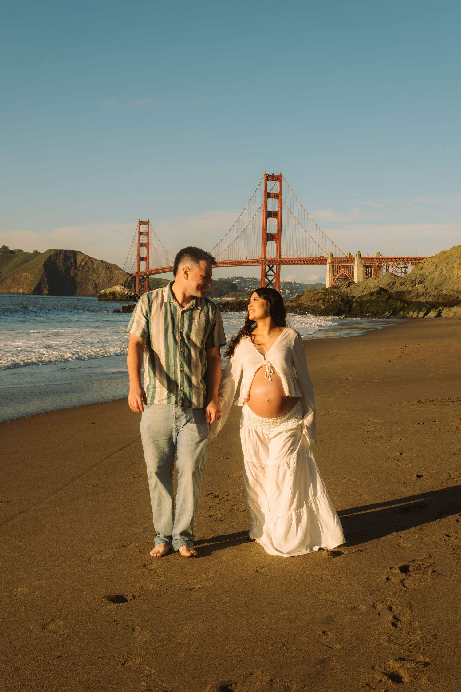 Pregnant couple walking on Baker Beach with Golden Gate Bridge backdrop in San Francisco at golden hour.