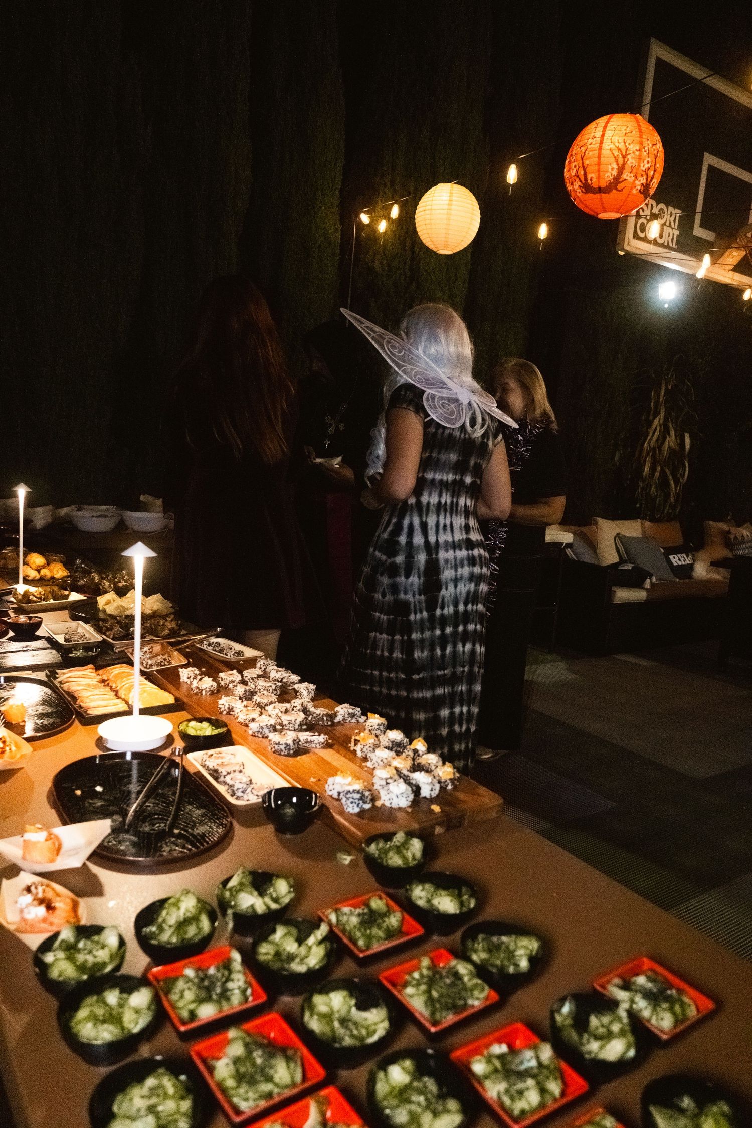 Halloween party buffet table with sushi and salads, woman in witch hat costume, paper lanterns glowing in dark background.