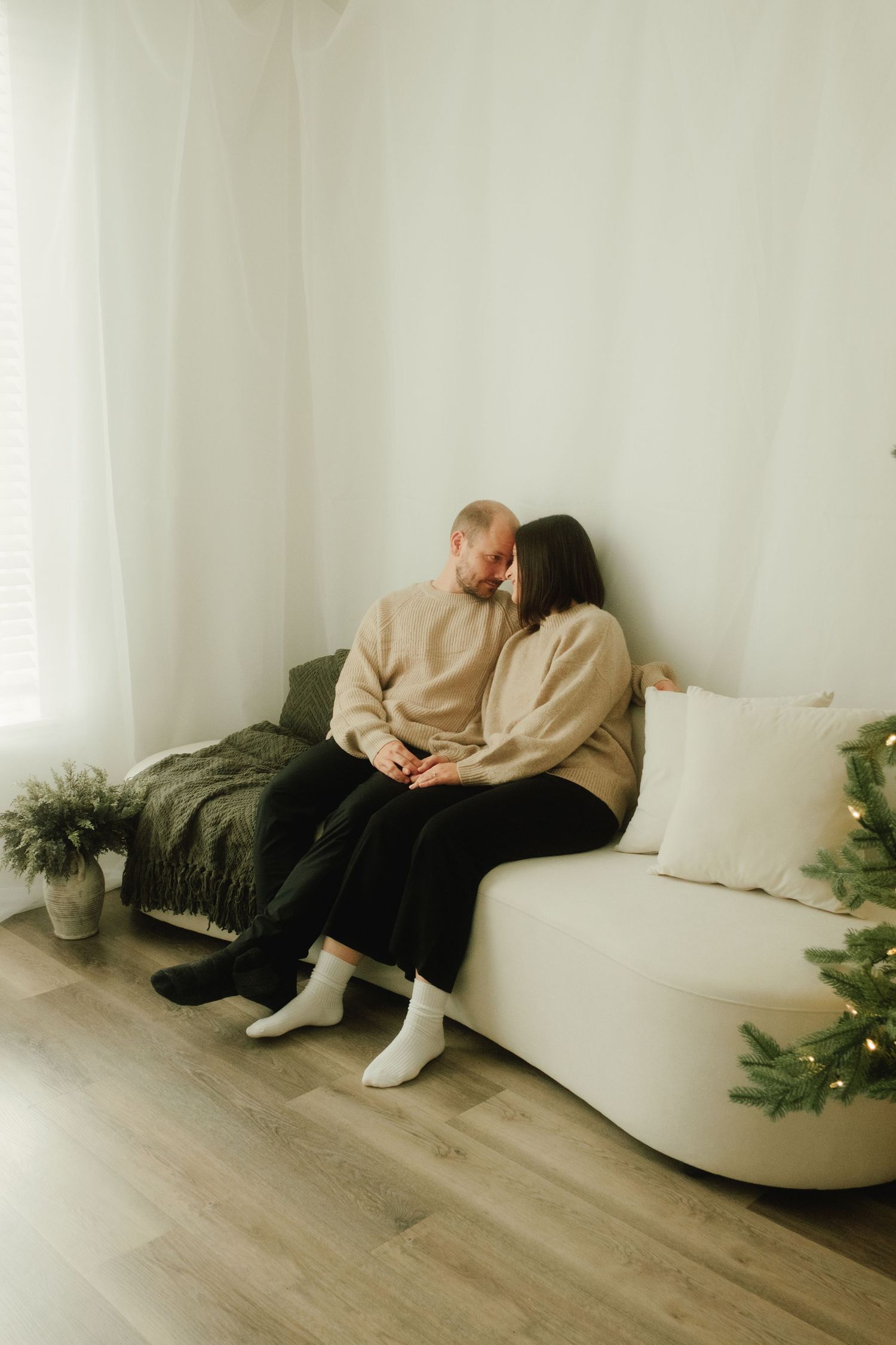 Couple in matching sweaters sitting close together on white couch with Christmas decor.