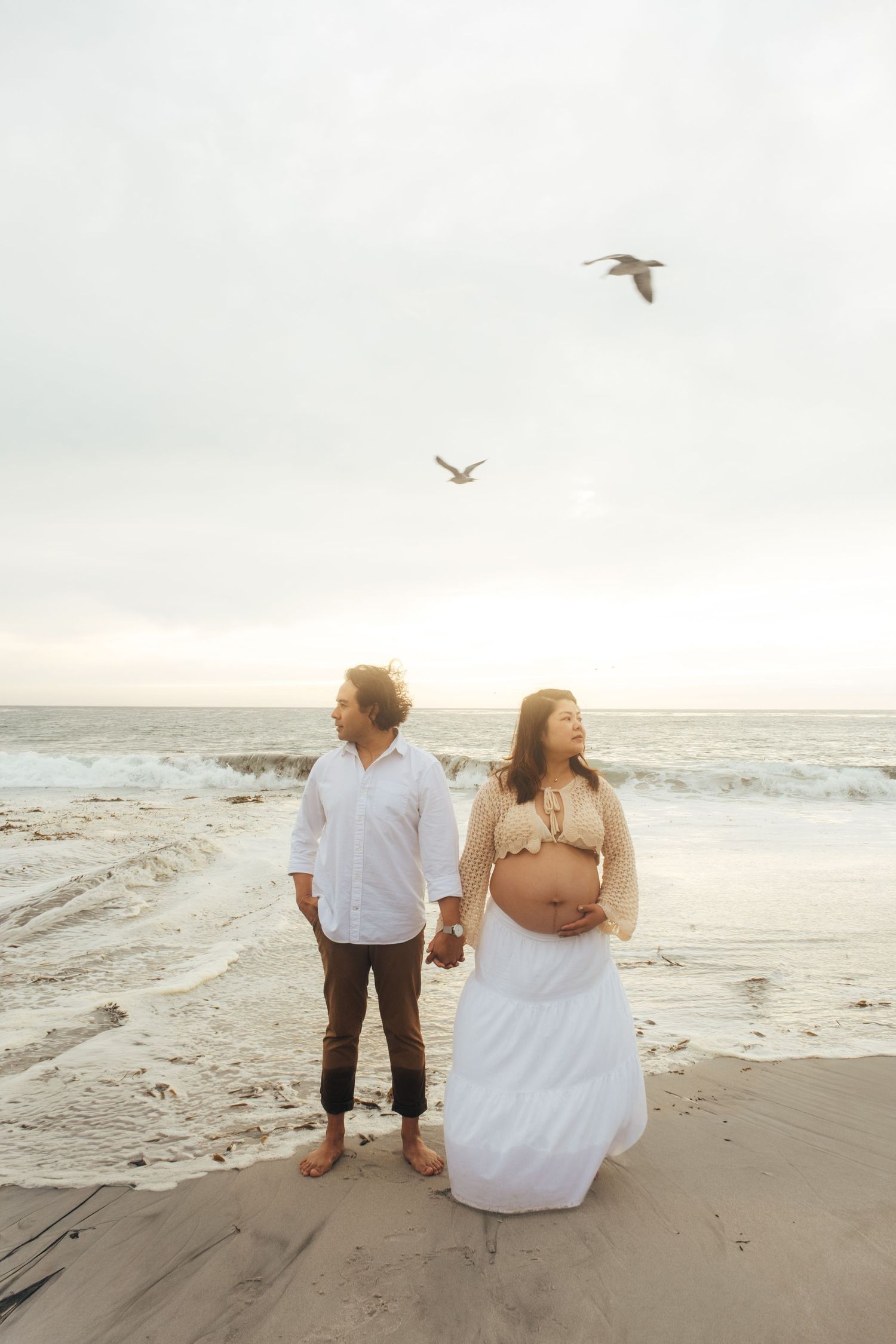 Couple walks along misty beach shore with seagulls flying overhead.