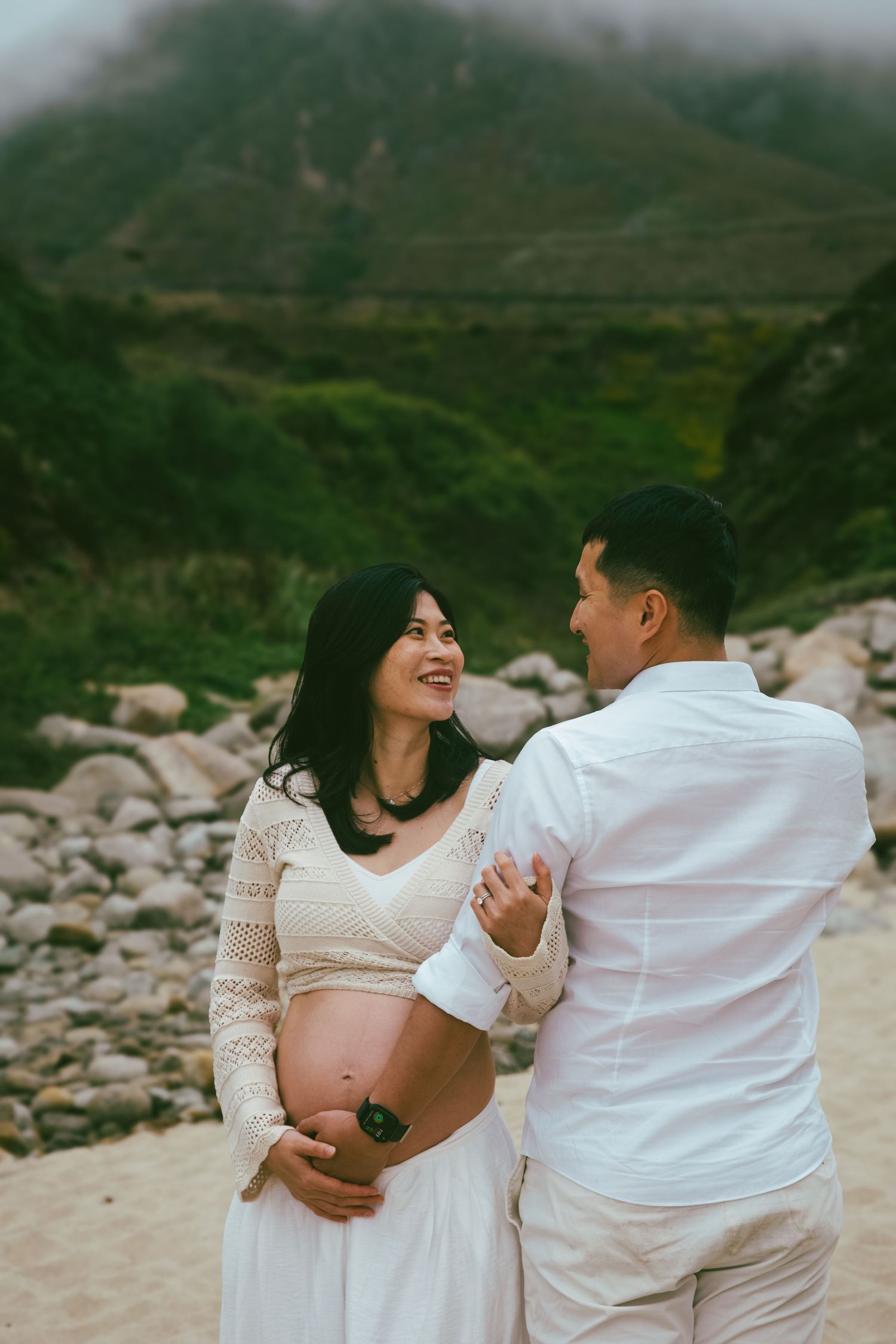 Couple in white outfits share intimate moments on a rocky beach during a romantic photoshoot.