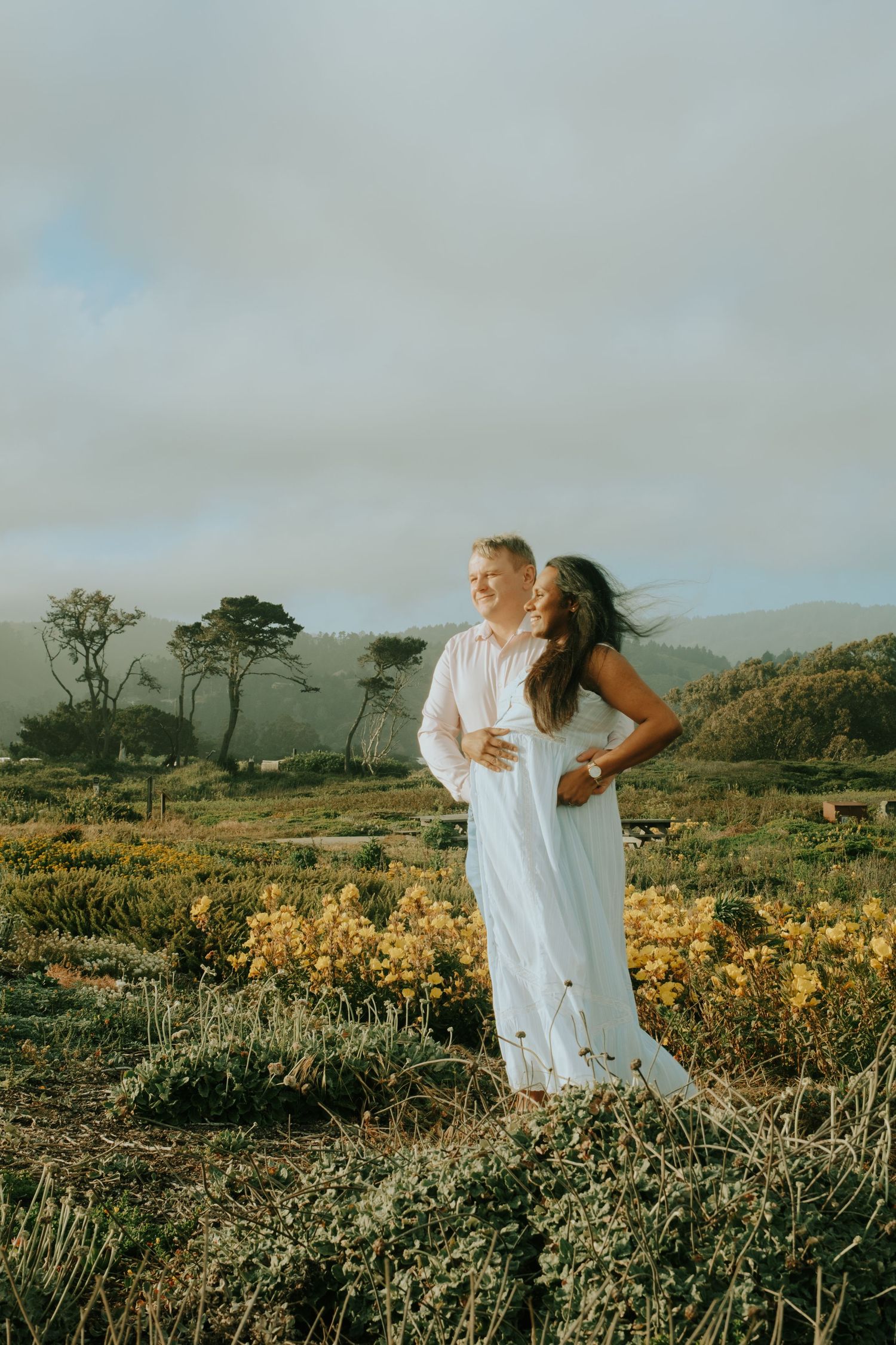 A couple poses in a field of yellow wildflowers with coastal scenery in the background.