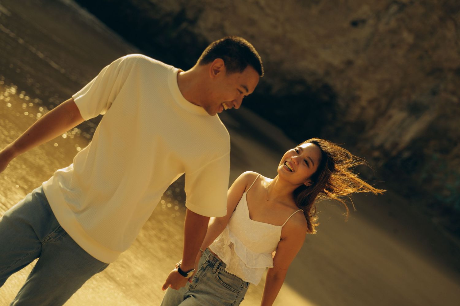 Happy couple holding hands and smiling on a sunny beach, golden light illuminating their casual summer outfits.