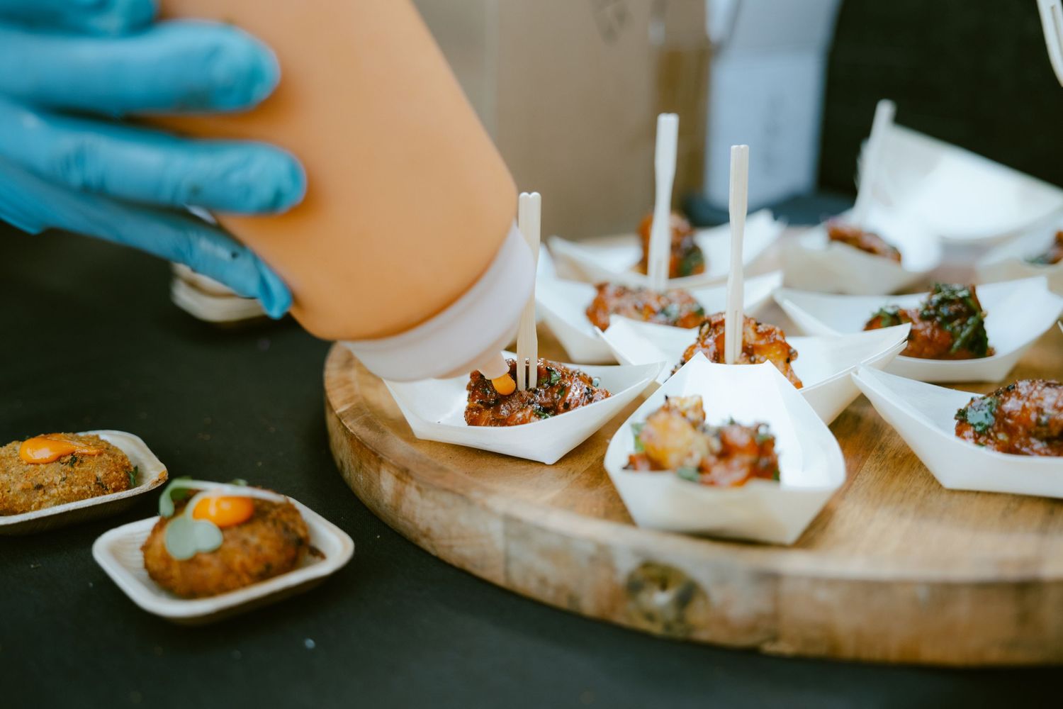 Gloved hand drizzling sauce over gourmet appetizers on a wooden board at a catered event.