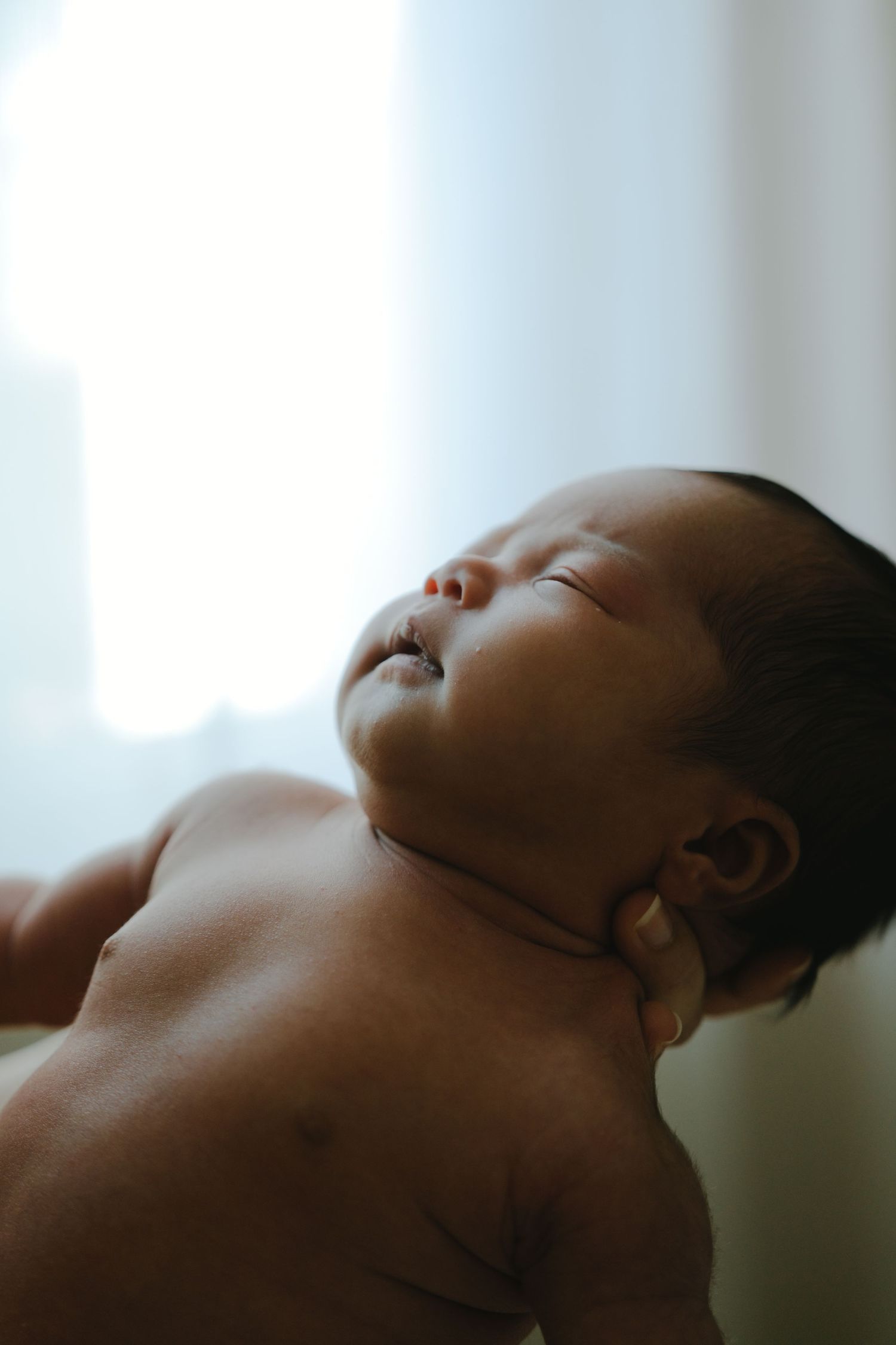 A peaceful newborn baby sleeps with eyes closed, wearing a small clip, in soft natural light.