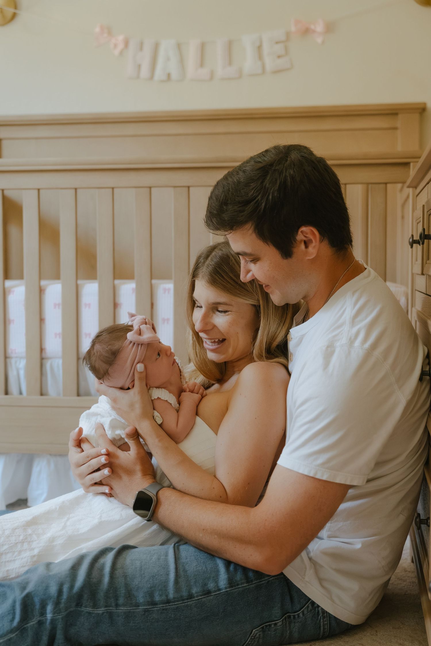 Happy parents sitting by a crib smiling at their newborn baby girl named Hallie in a soft nursery.