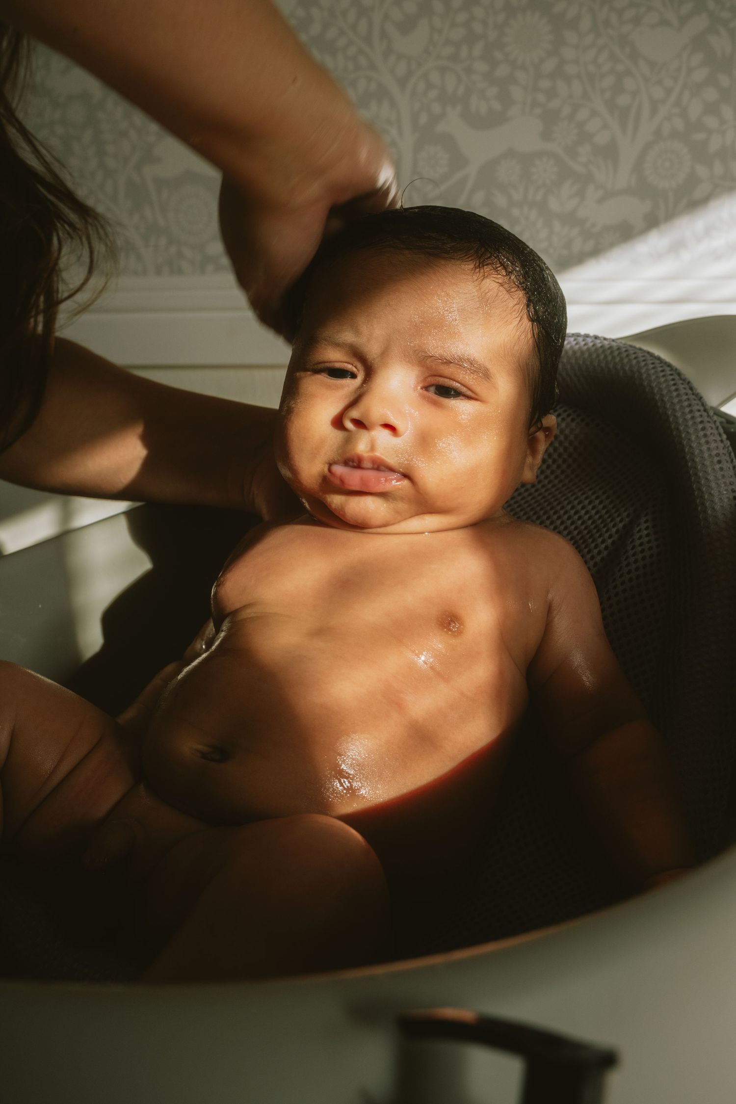 Newborn baby enjoying first bath in round tub, supported by parent's hands in warm sunlit room.