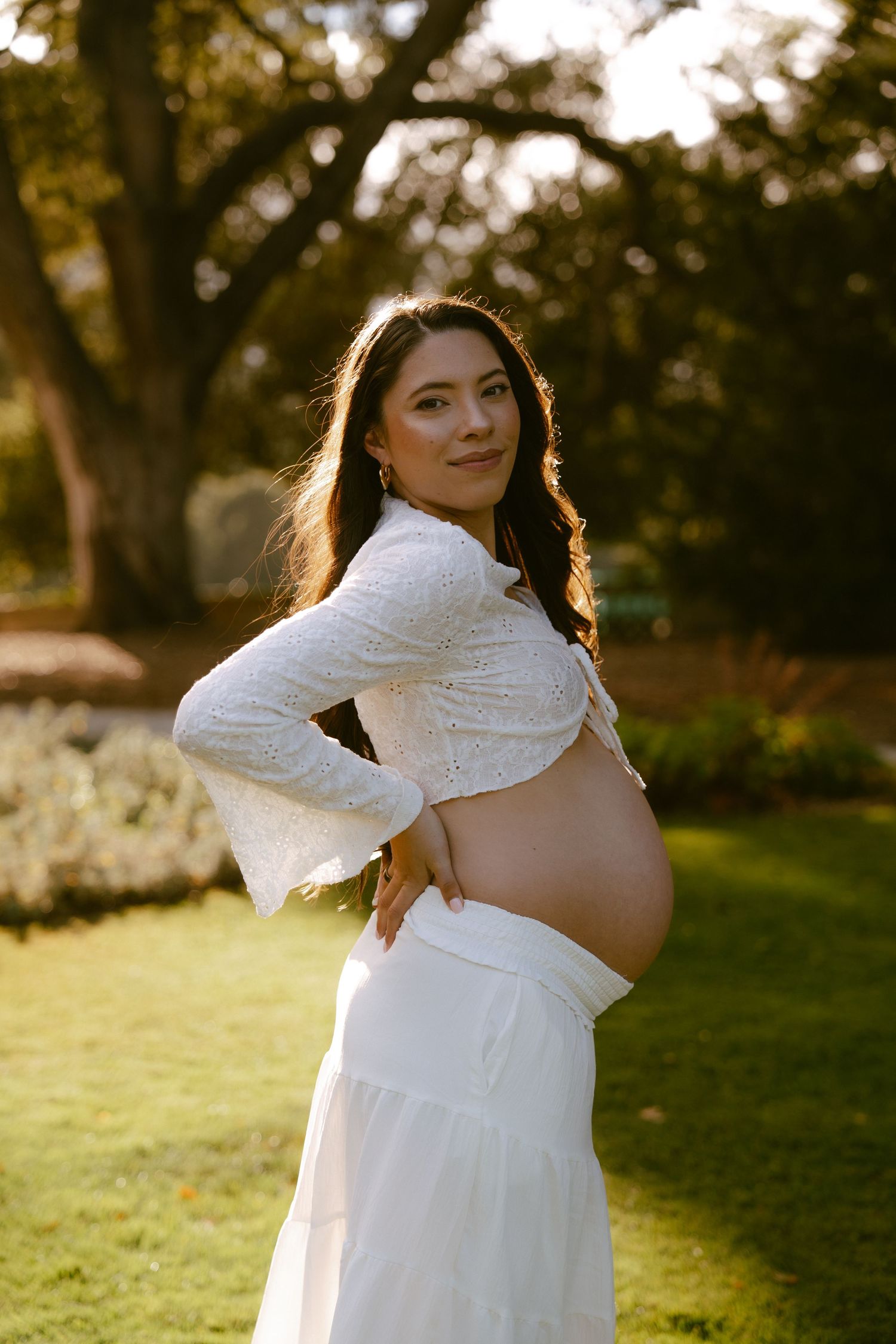 Pregnant woman in white crop top and skirt posing outdoors in golden hour sunlight with lush green trees behind her.