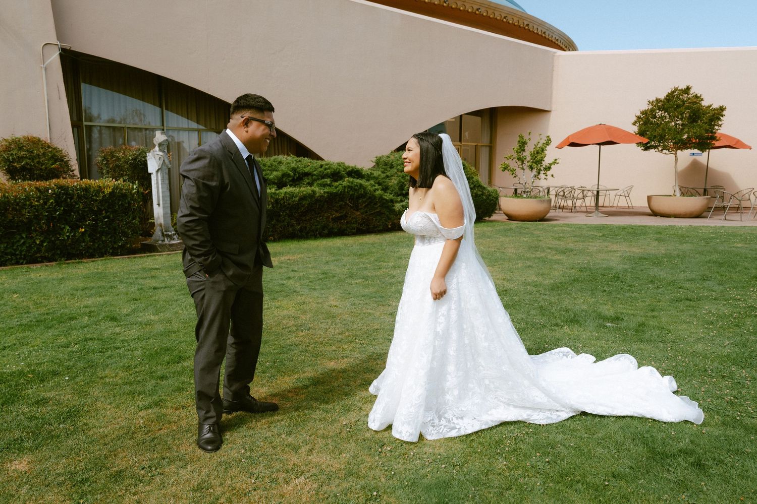 Bride in white gown and groom in black suit share a first look moment on a green lawn outside a venue.