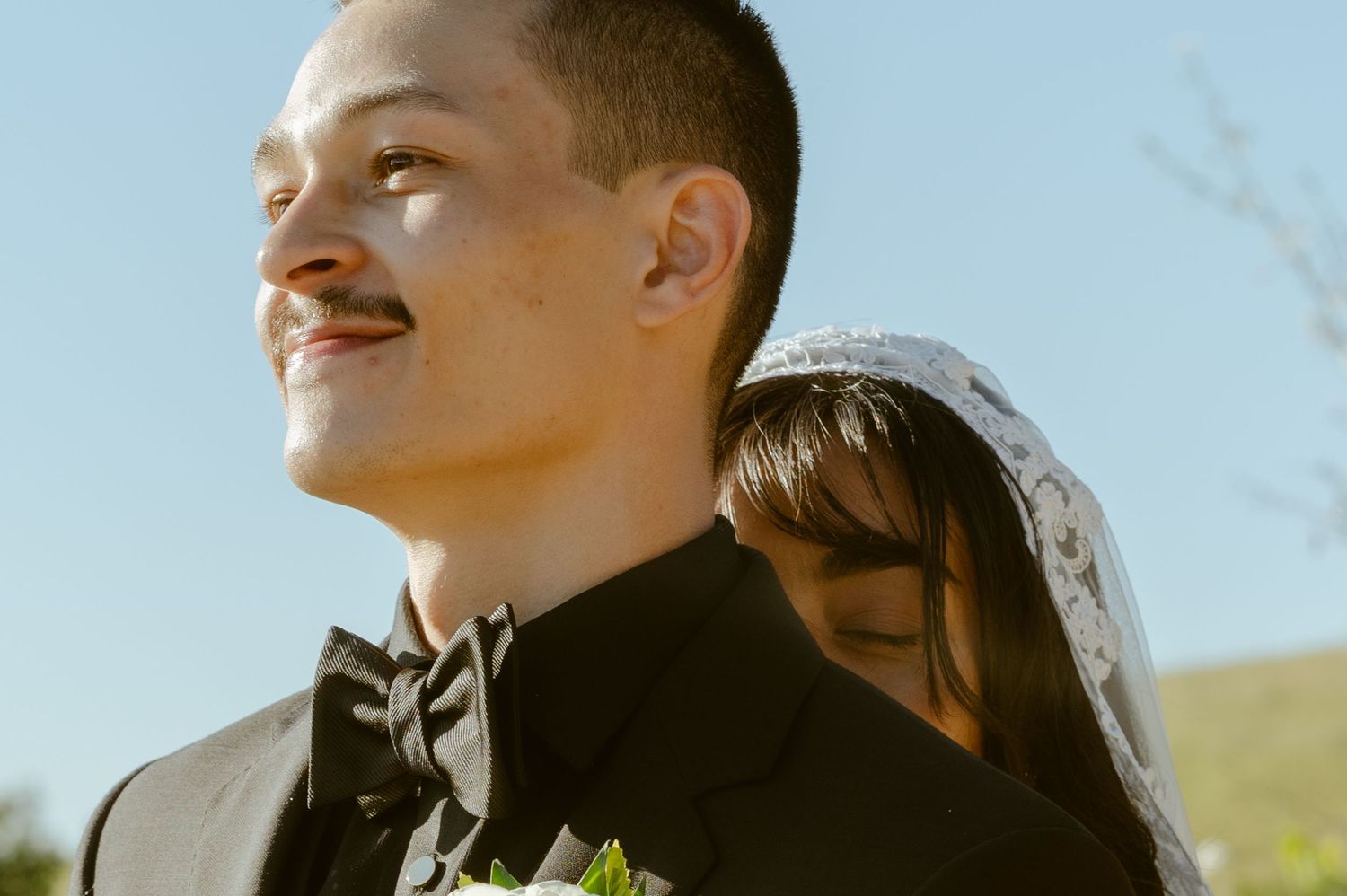 A smiling groom in a black tuxedo with boutonniere stands outdoors as bride in white veil leans behind him.