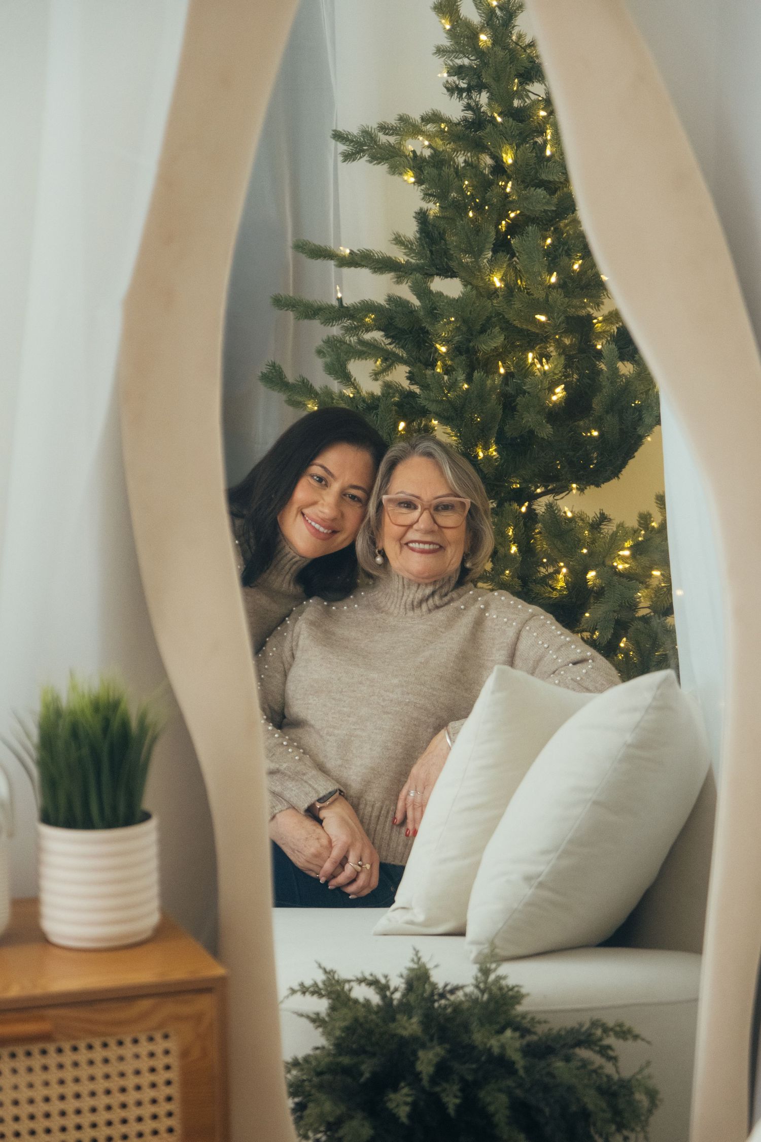 Two people pose together in cozy nook with Christmas tree and home decor visible in background.