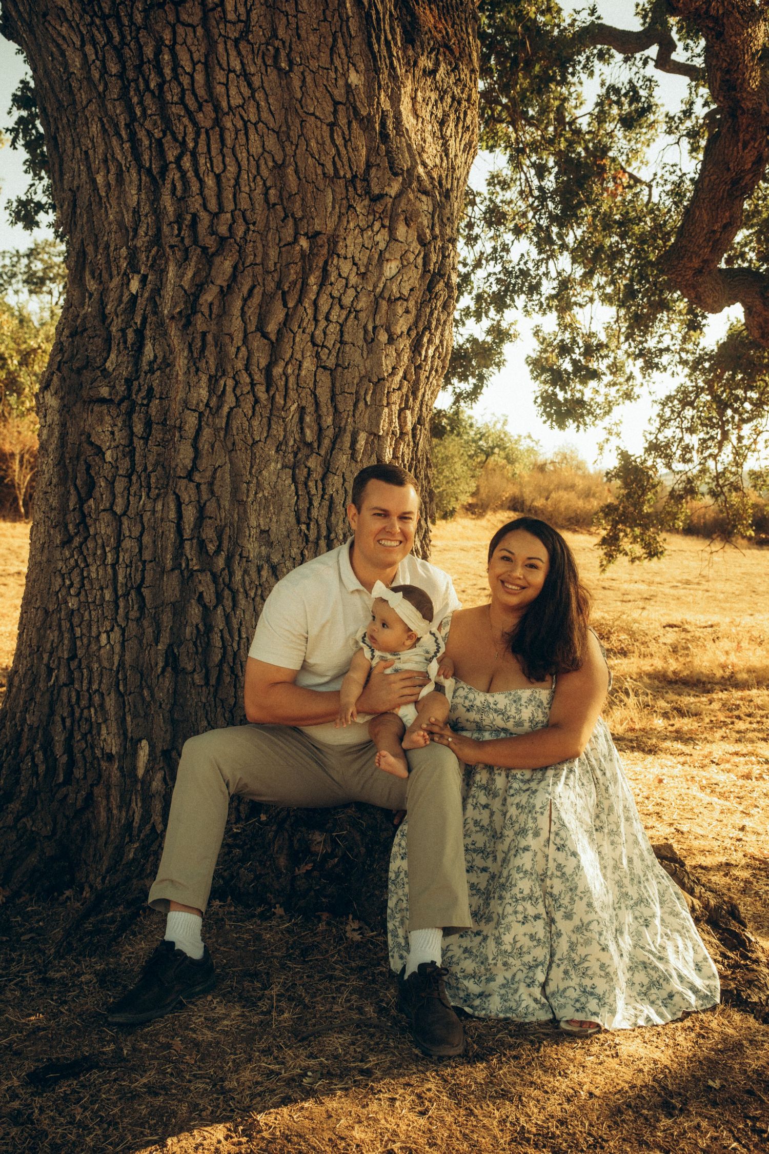 A couple sits together under a large oak tree in golden afternoon sunlight.