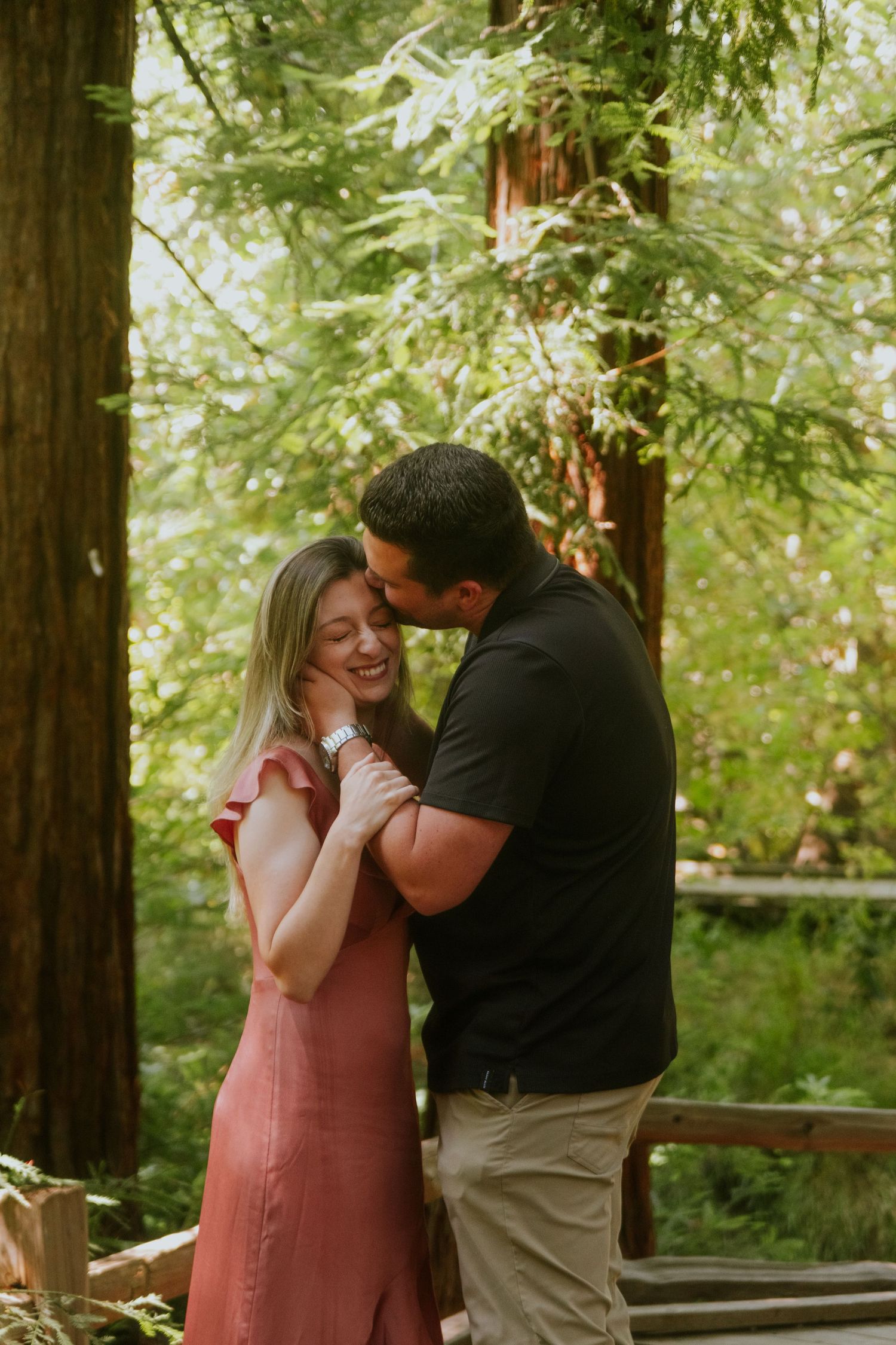 A couple embraces tenderly on a forest boardwalk surrounded by towering redwood trees.