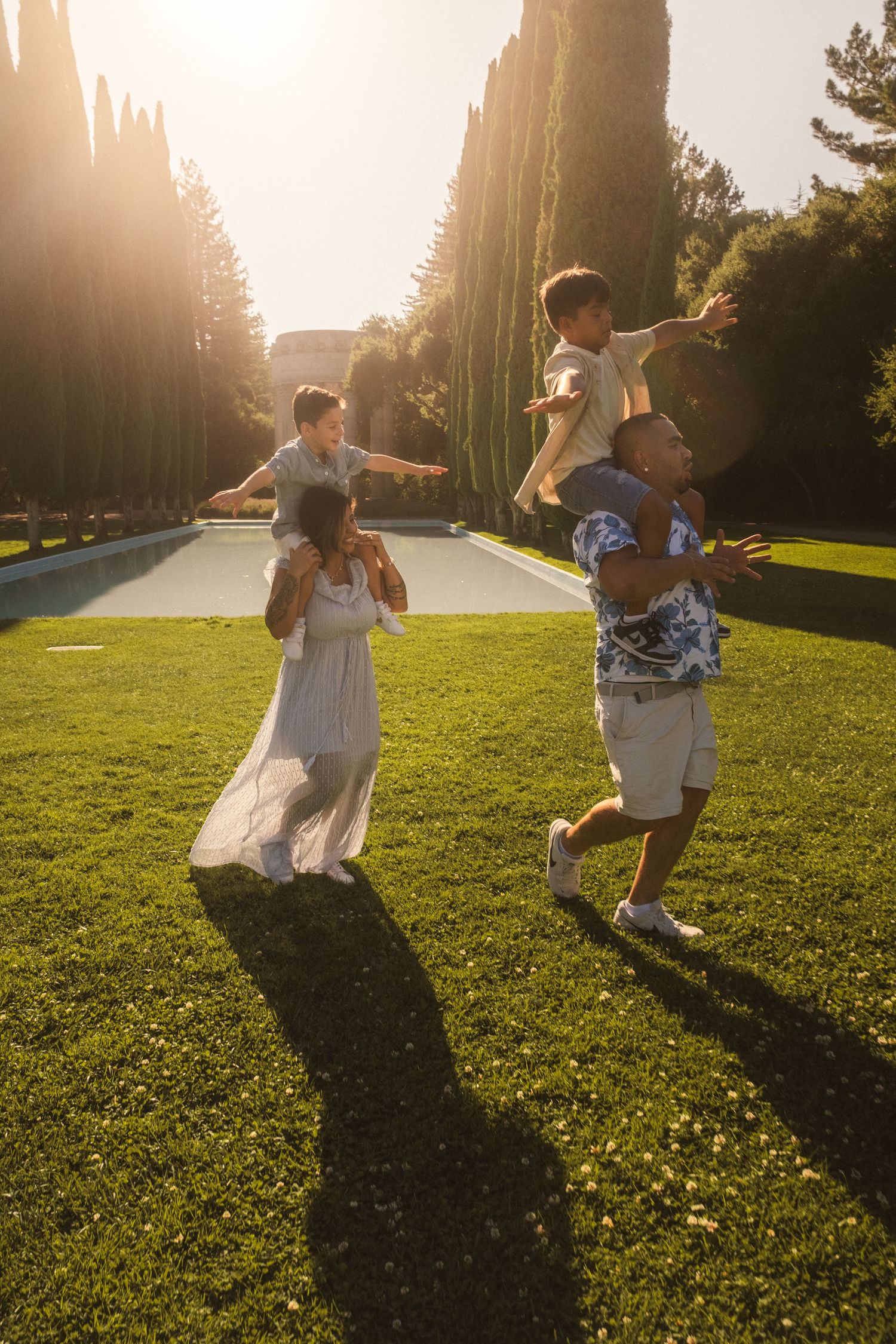 People playing and running joyfully across a sunlit grassy field with cypress trees in background.