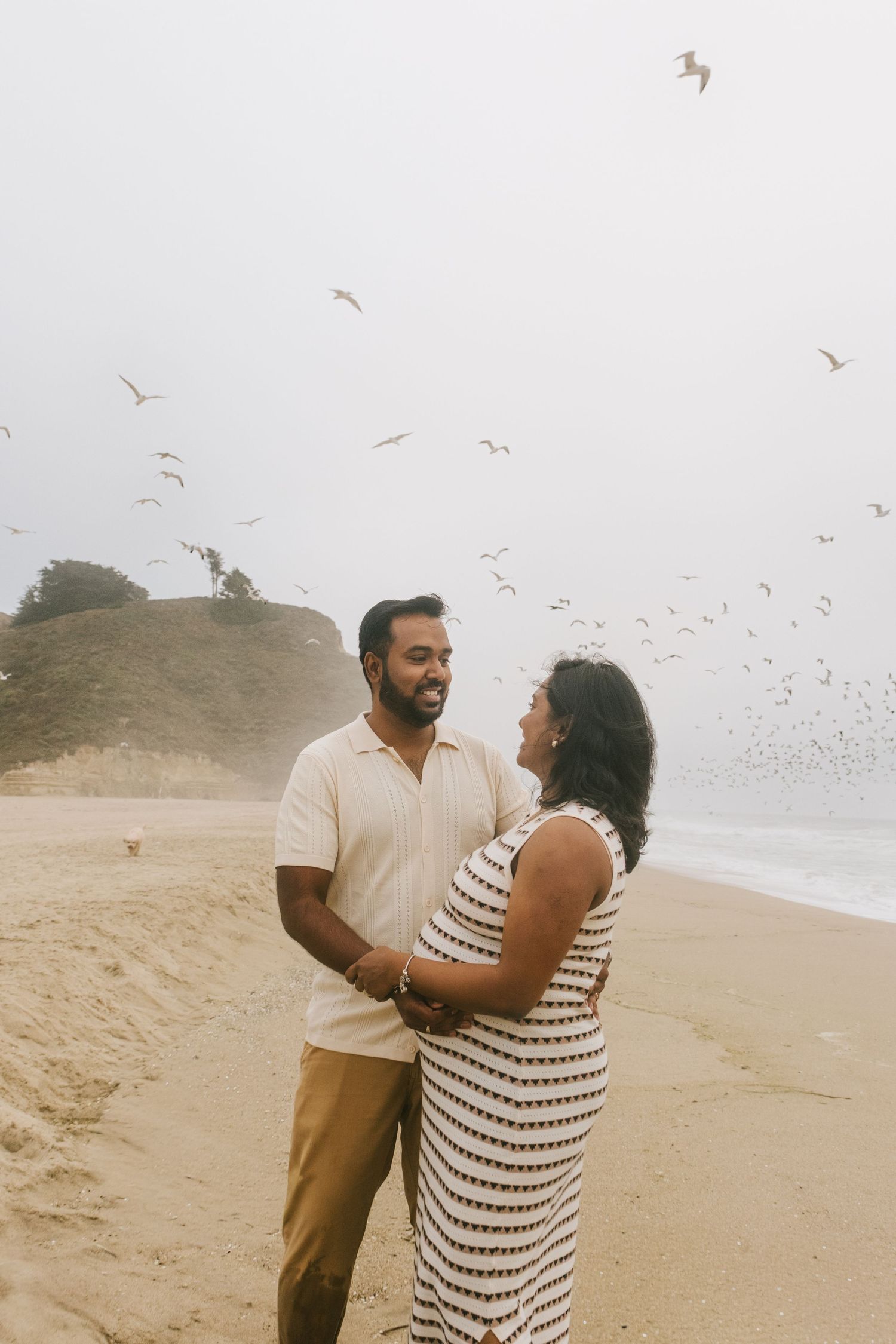 A couple poses on a beach with seabirds flying in the background.