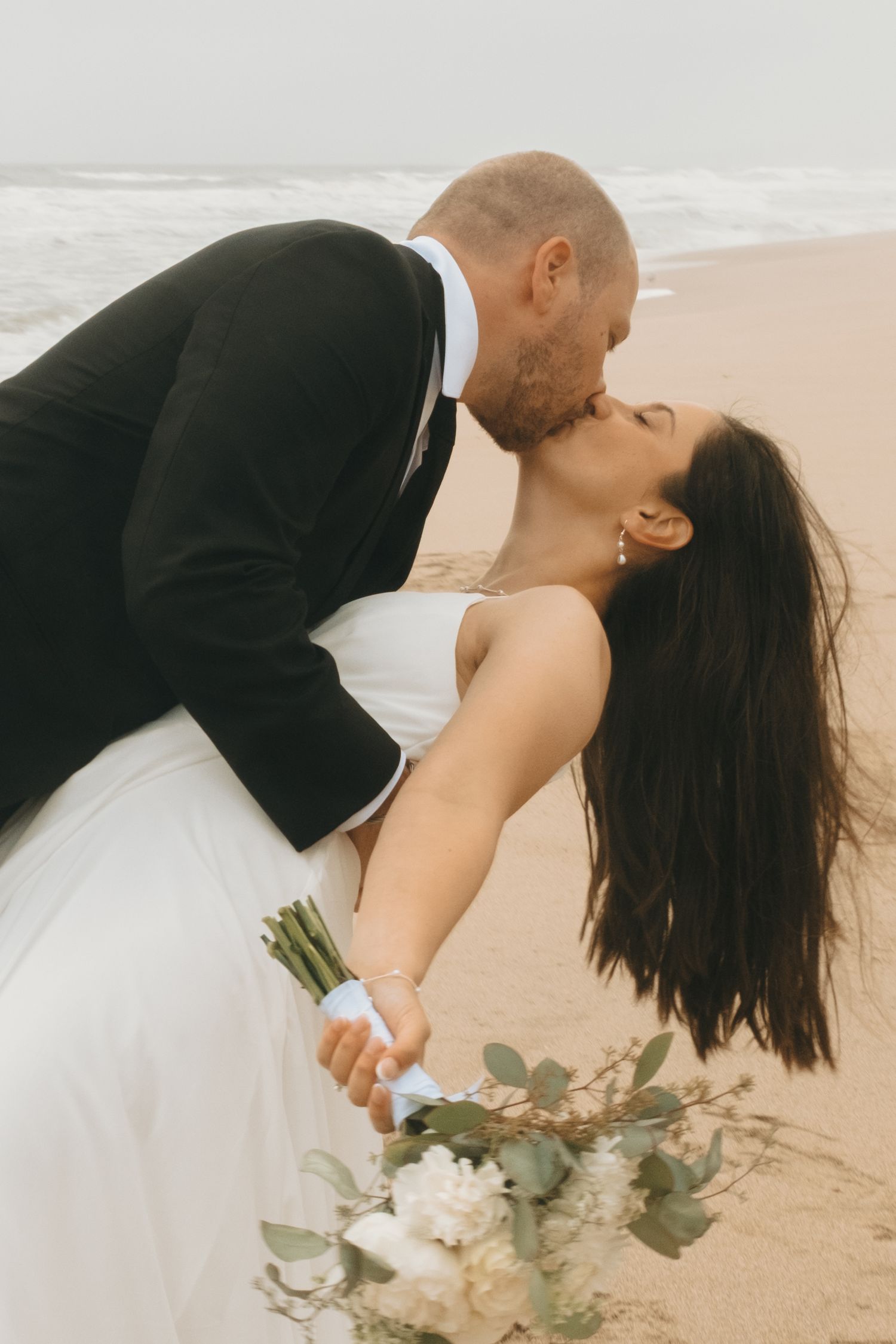A romantic beach wedding photo with the groom dipping his bride while holding a bouquet.