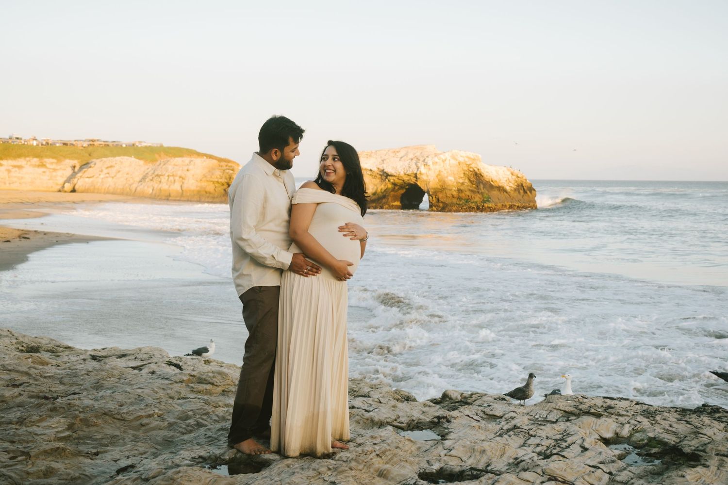 Romantic coastal photoshoot with dramatic rock formations and ocean waves in background.