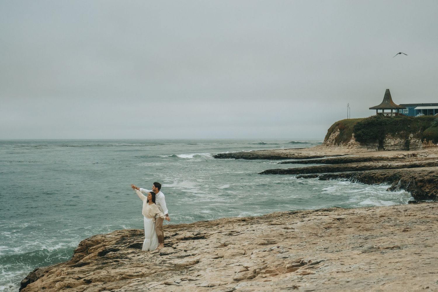 Romantic coastal photoshoot at a scenic beach with rocky cliffs and crashing waves during golden hour.