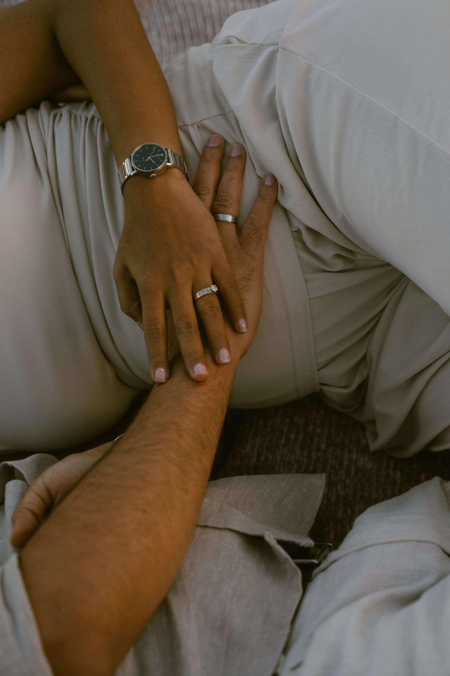 Close-up of couple's hands with wedding rings and a silver watch, resting on light gray fabric.