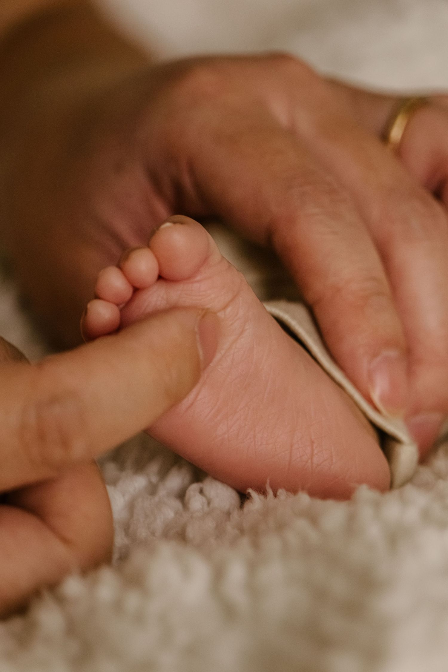Adult hands tenderly holding tiny newborn baby feet on a soft white blanket, with a gold ring visible.