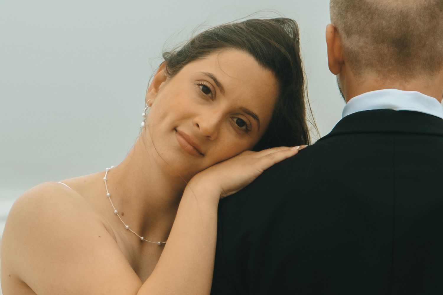 Bride in pearl necklace rests her head on groom's shoulder, gazing up lovingly during wedding ceremony.