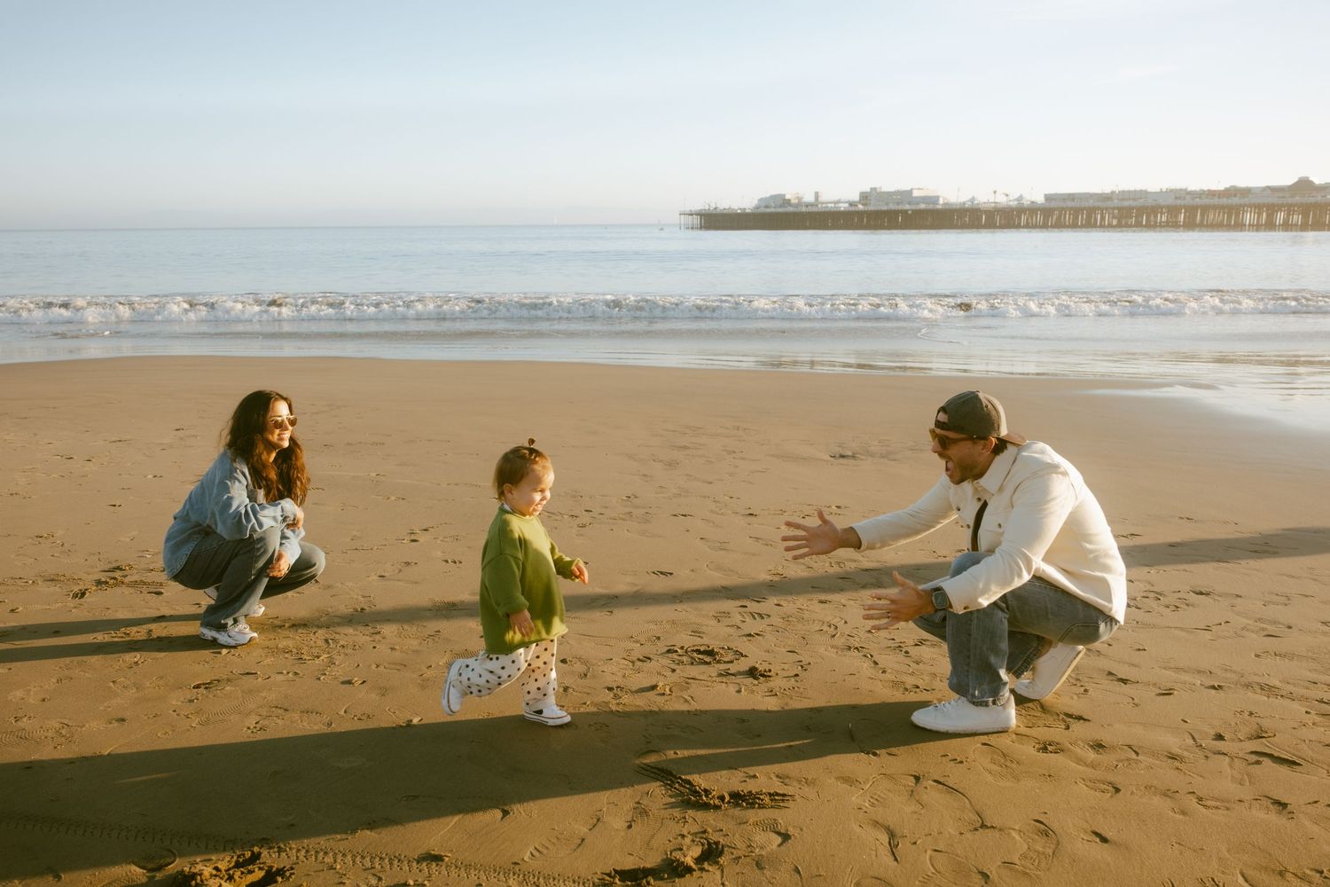 Family enjoying a sunny beach day, with parents crouching as toddler walks between them near a pier.