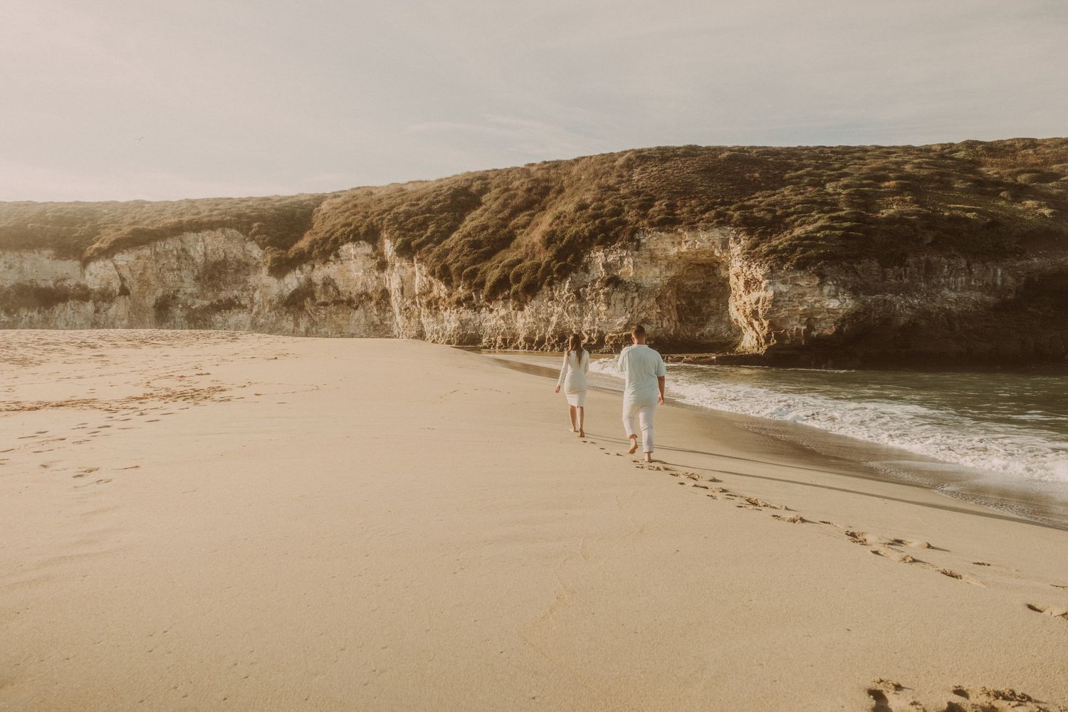 Couple walking hand in hand along a sandy beach with dramatic cliffs in the background.