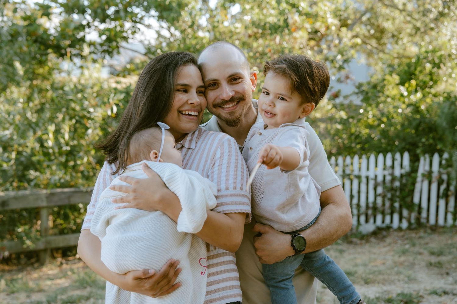 Happy family of four posing outdoors, parents holding two young children, smiling in a lush garden setting.