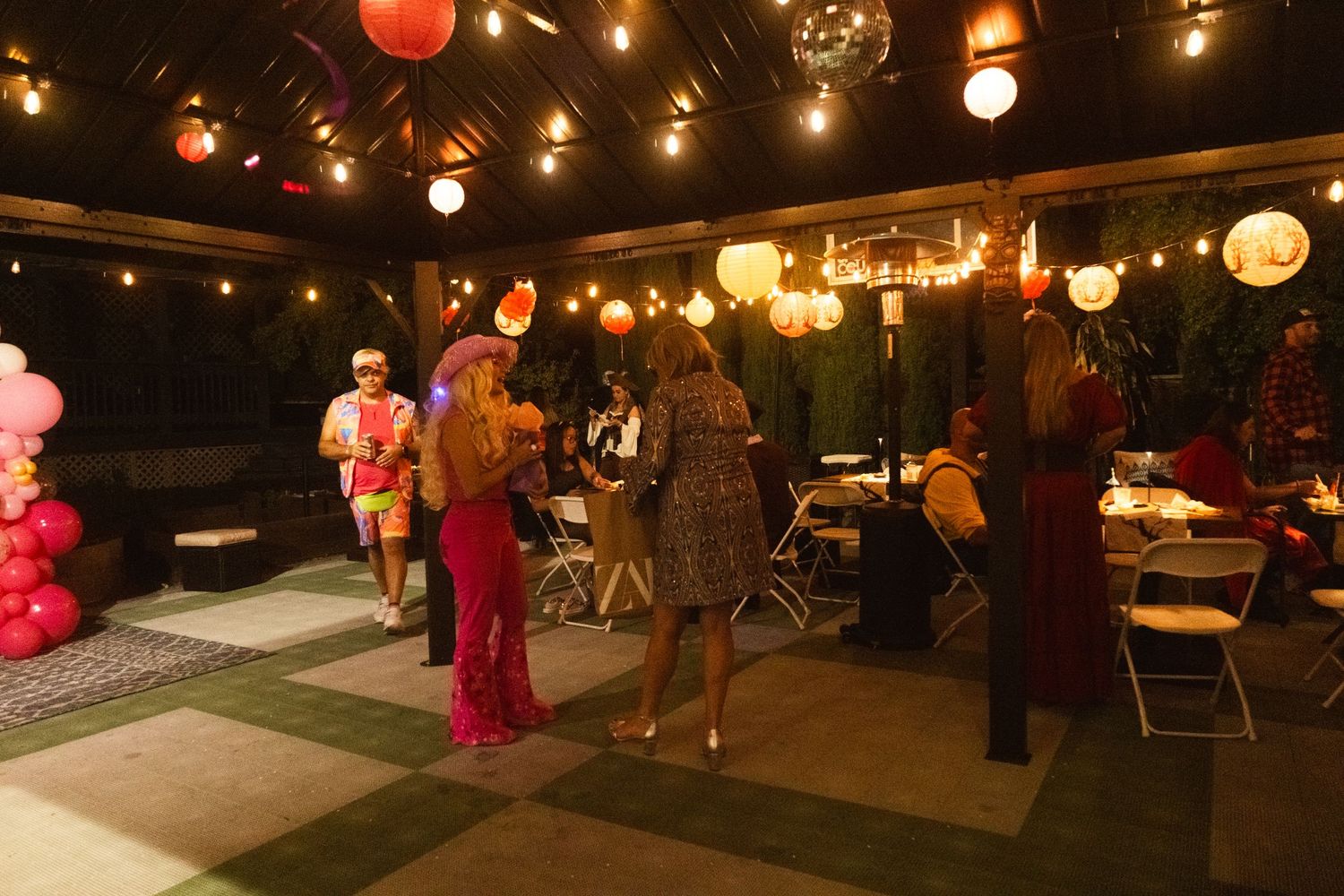 Guests mingle at an outdoor evening party with string lights and colorful lanterns decorating the festive venue.