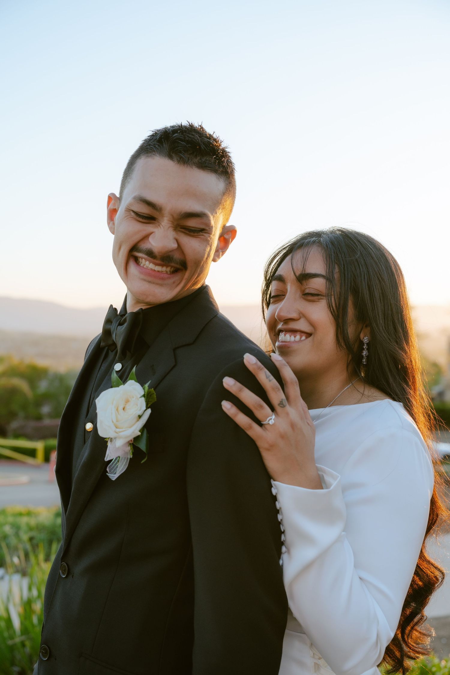 Smiling newlywed couple posing outdoors at sunset, groom in black suit with white boutonniere, bride showing wedding ring.
