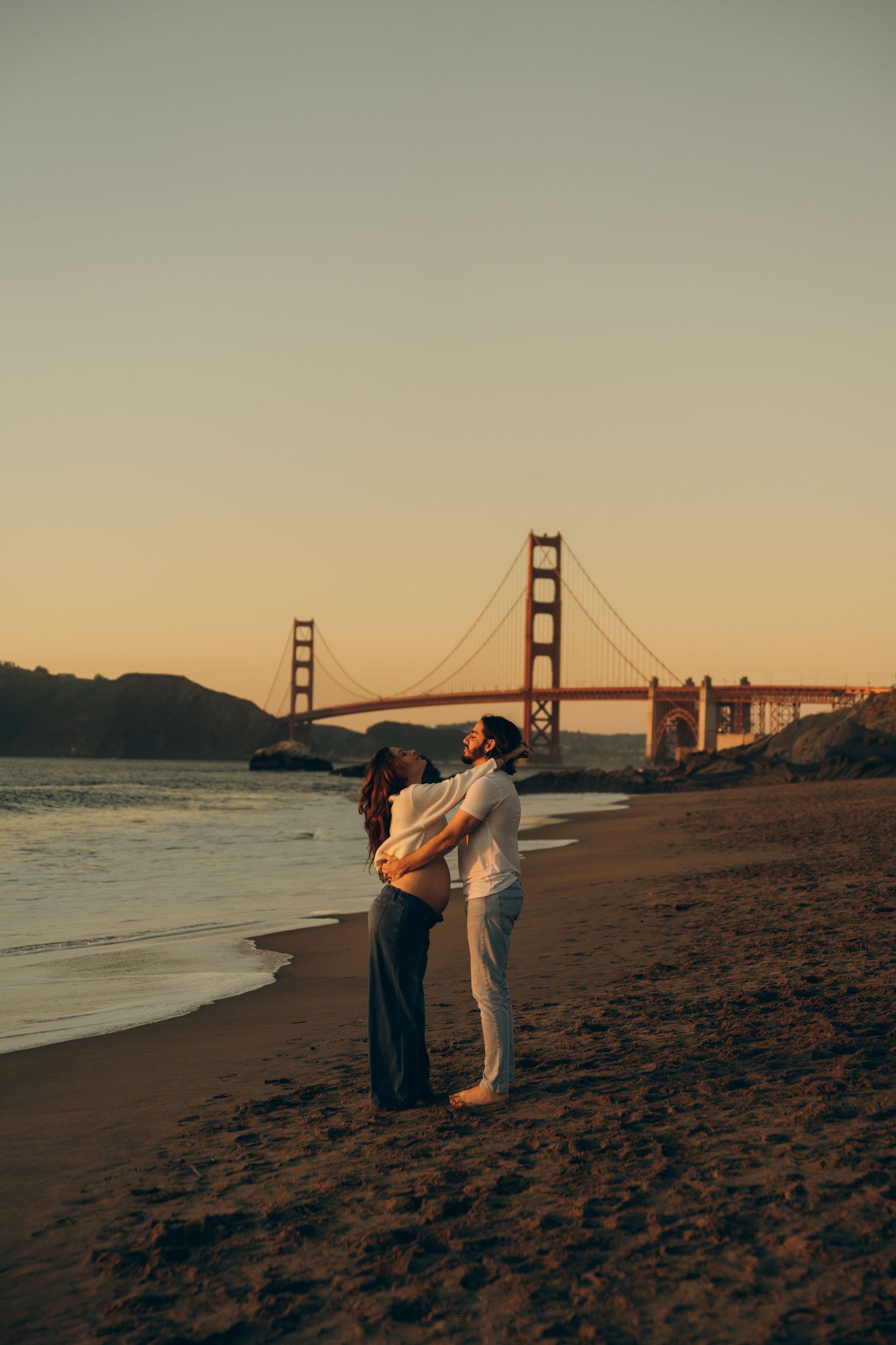 Couple embracing on Baker Beach at golden hour with the iconic Golden Gate Bridge glowing in the background.