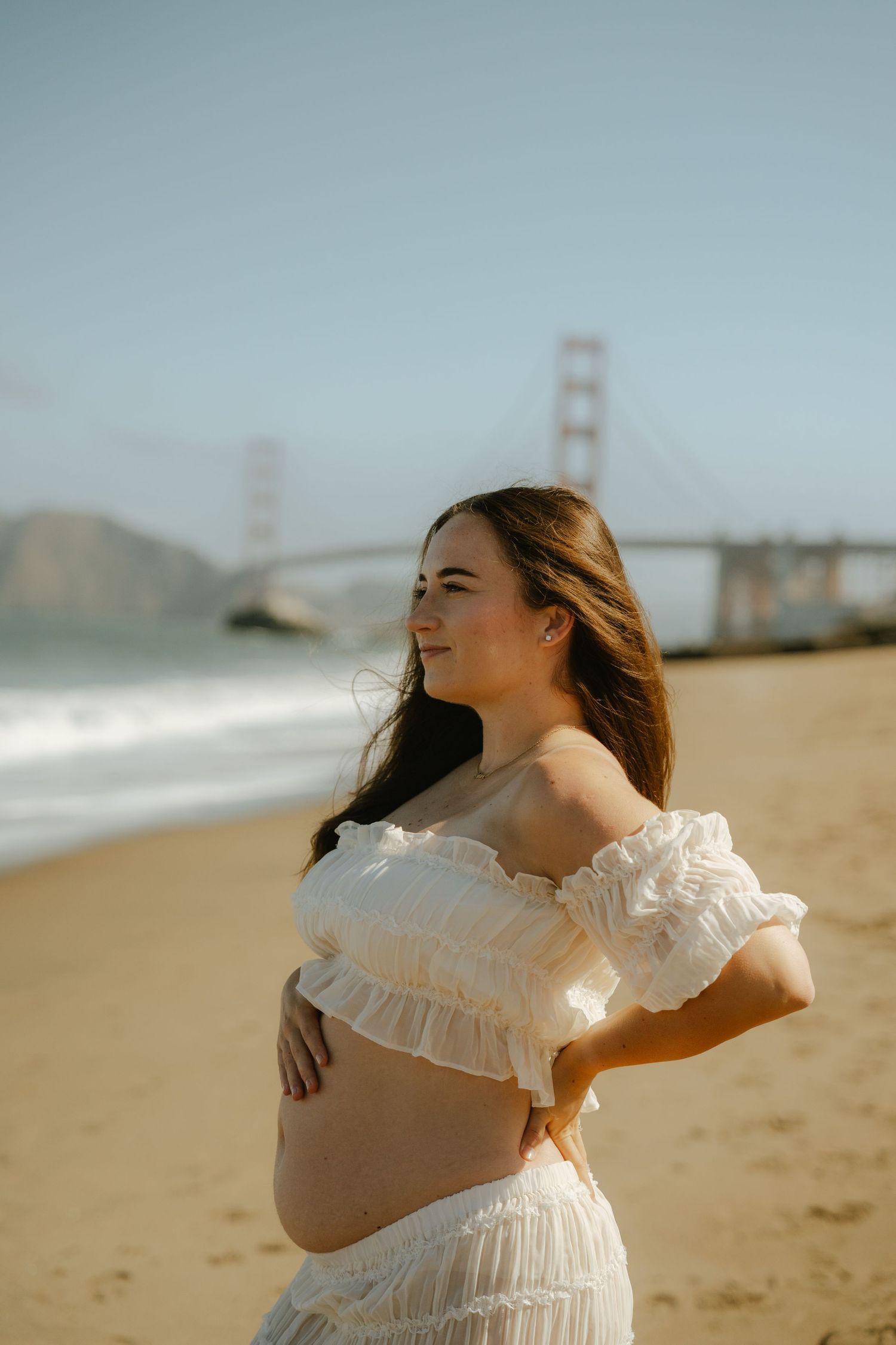 Pregnant woman in white off-shoulder dress cradling belly on sandy beach with Golden Gate Bridge in background.