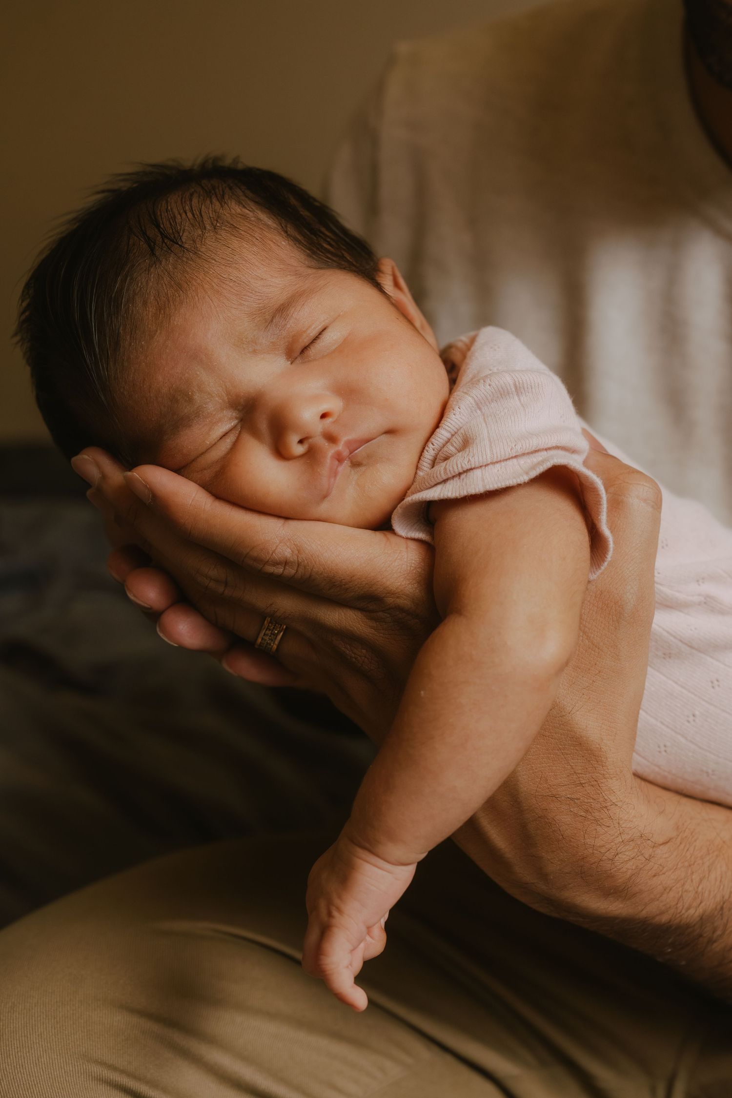 Newborn baby sleeping peacefully in parent's hands, wearing a pink onesie, with soft warm lighting.