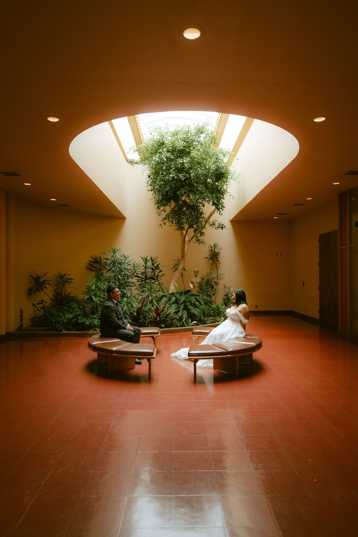 Bride and groom sit on circular benches beneath a skylit atrium with lush tropical plants in an elegant indoor setting.