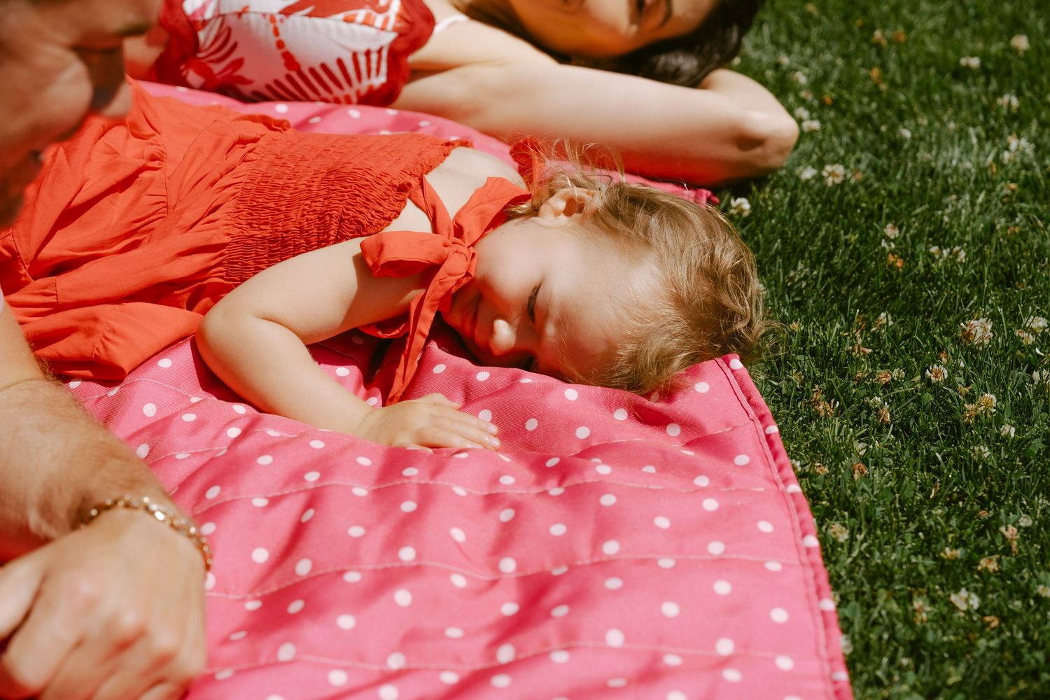 Baby in red dress sleeping on pink polka dot blanket outdoors on grass with family nearby.
