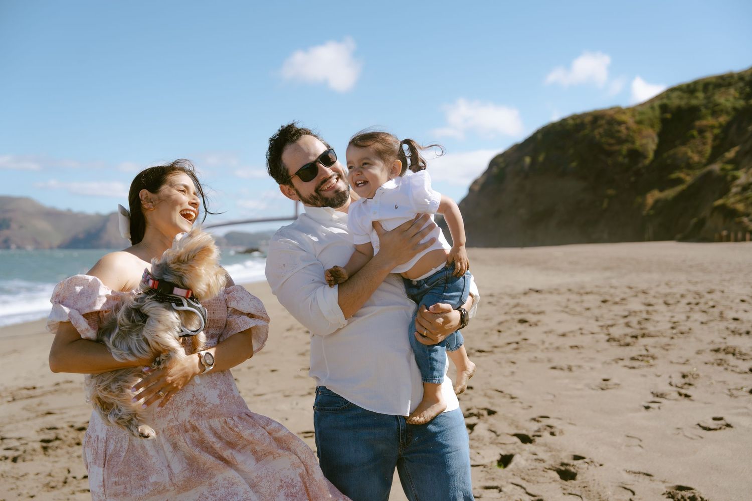 Happy family with two young children and a dog laughing together on a sunny beach with scenic cliffs.