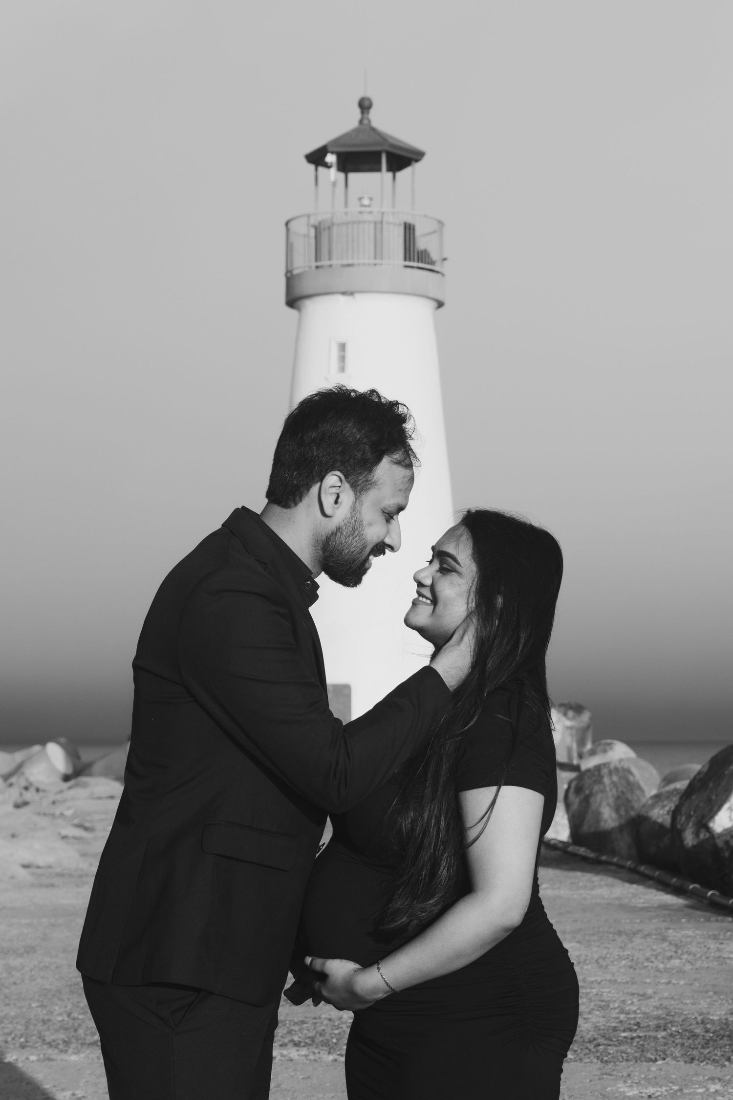 A couple poses romantically near a lighthouse in black and white photography.