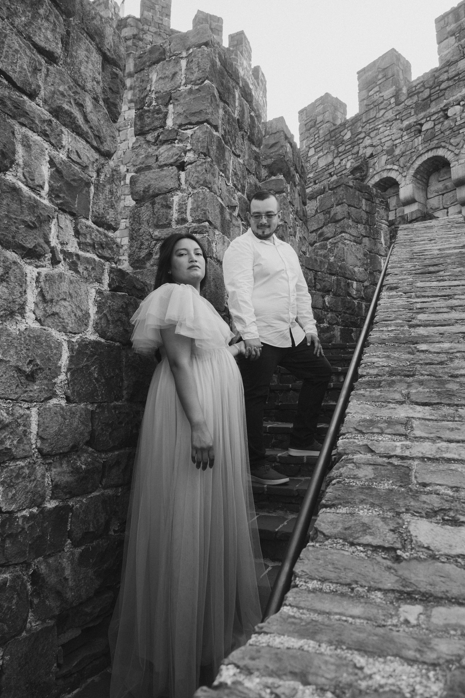 A couple poses on stone steps against medieval castle walls in black and white photography.