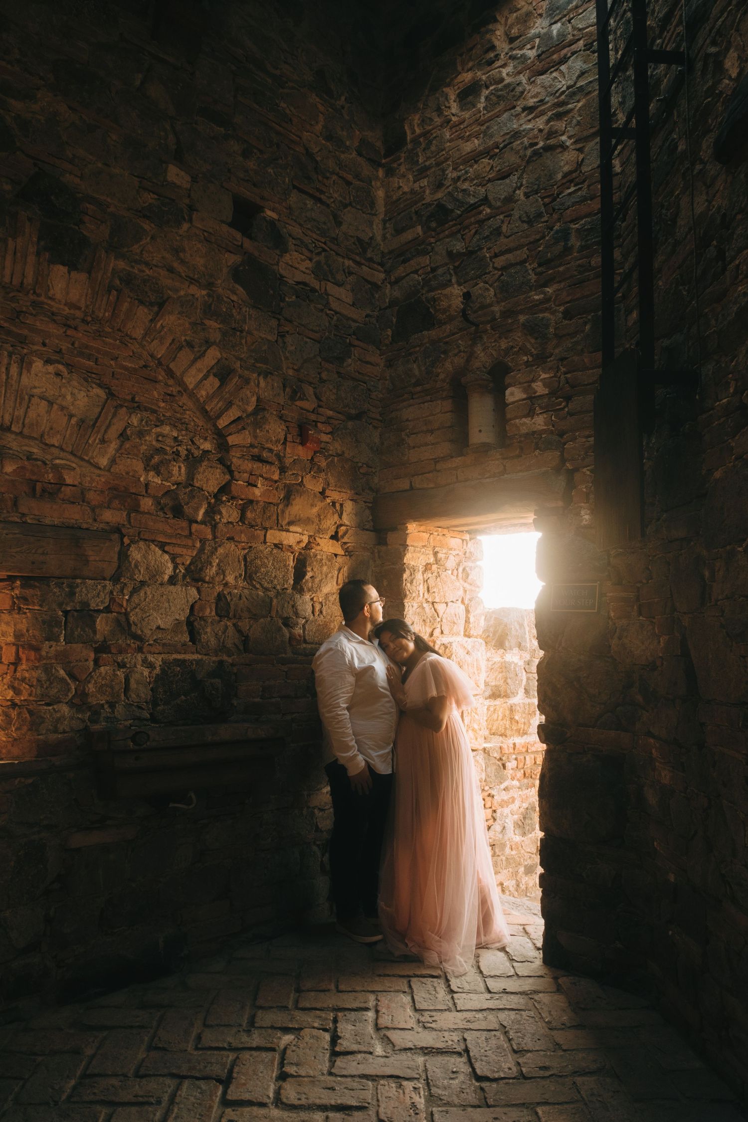 A couple stands in dramatic lighting within an ancient stone passageway.
