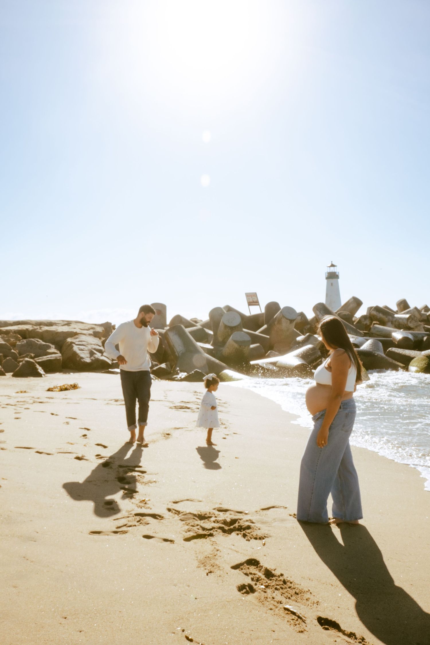 Family enjoying sunny day at beach with lighthouse and rocky shore in background.