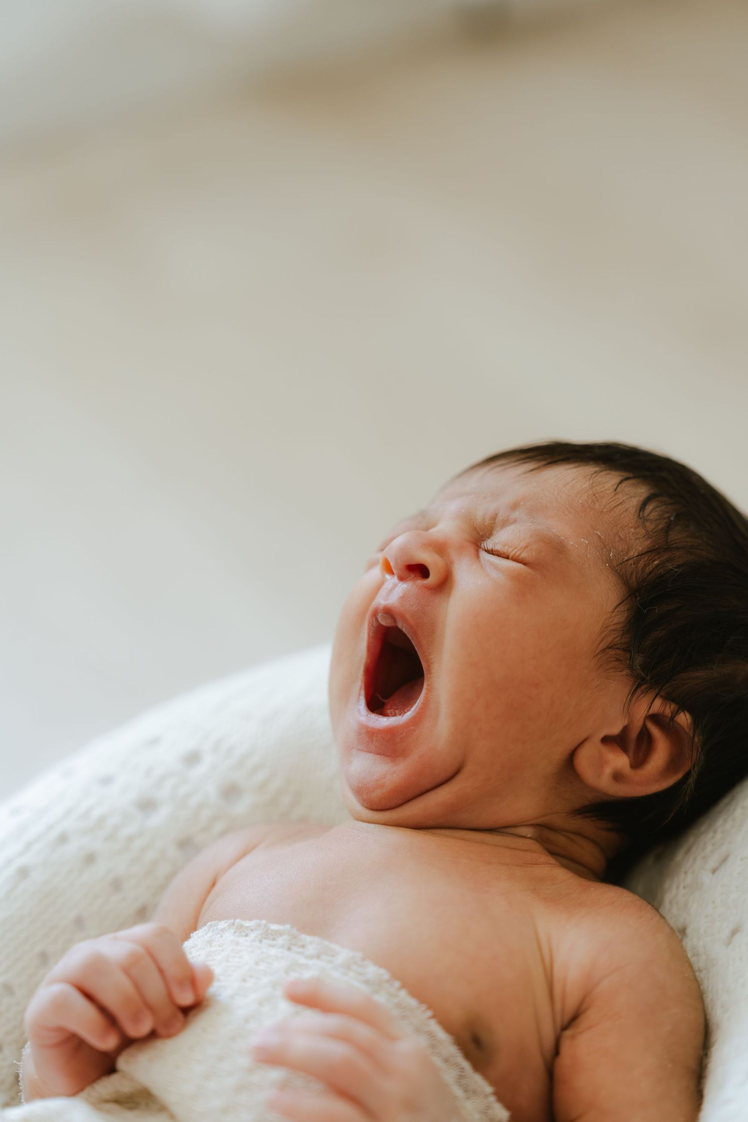Newborn baby yawning with mouth wide open, lying on a soft white blanket in a cozy basket.
