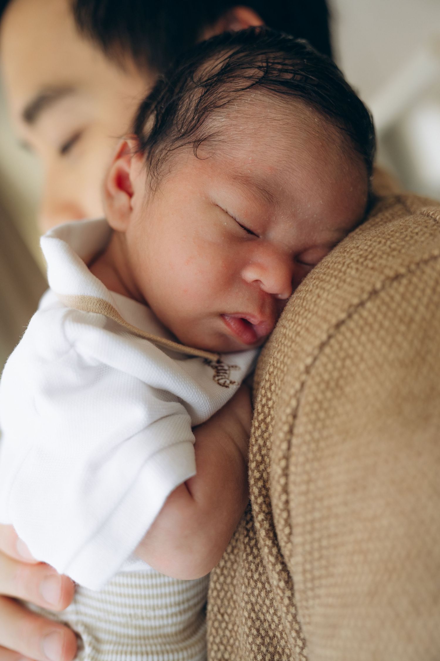 A newborn baby sleeping peacefully on a parent's shoulder, wrapped in a white onesie and striped pants.