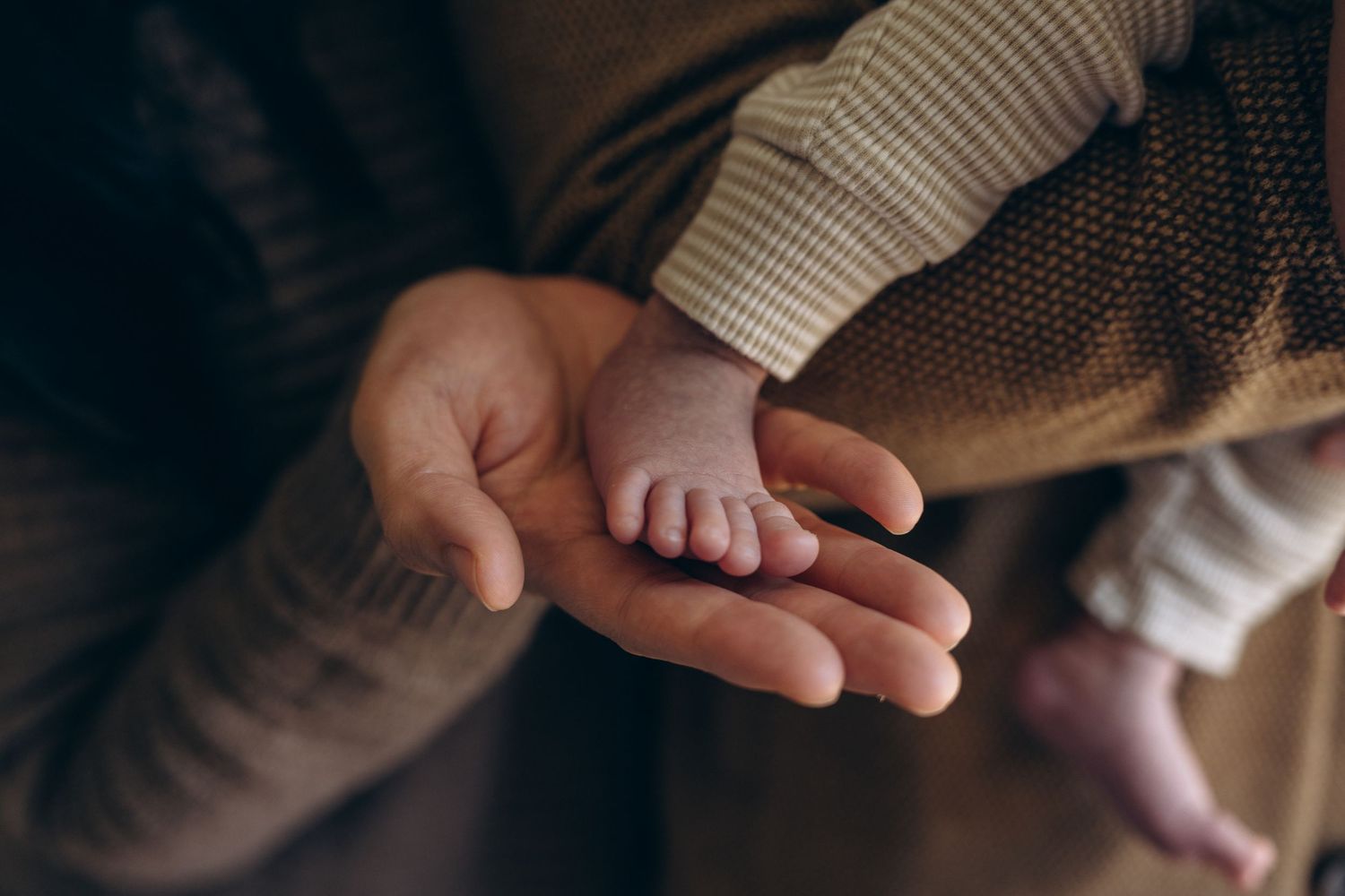 Adult gently cradling a tiny newborn baby foot in their open palm, showcasing a tender parent-child bonding moment.