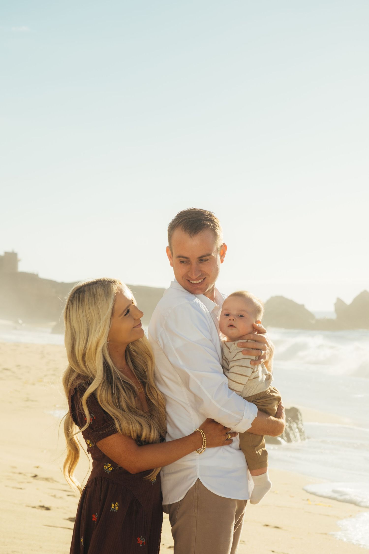 A family shares a tender moment on a sunny beach with rocky cliffs in the background.
