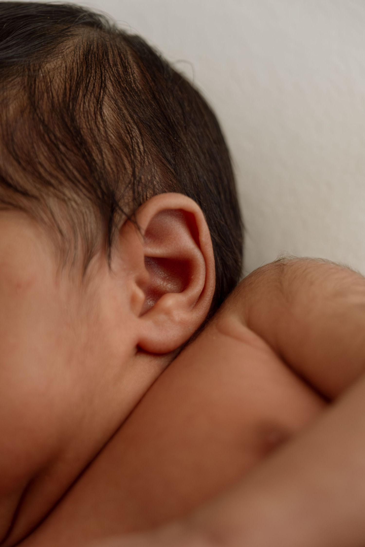 Close-up view of a baby's ear and dark hair against a neutral background.