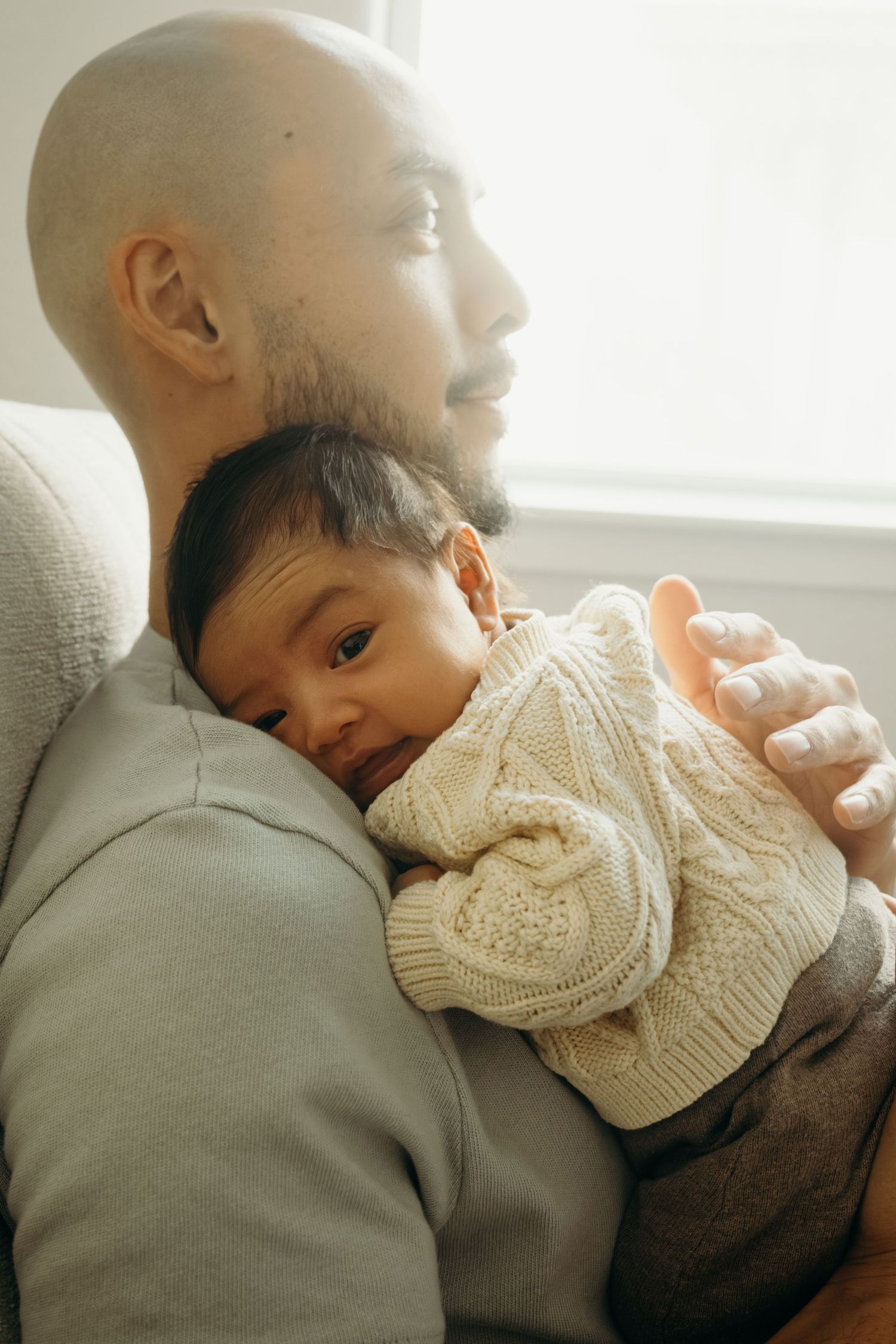 A bald bearded man tenderly holds a sleeping baby in a cream knit sweater against his chest near a bright window.